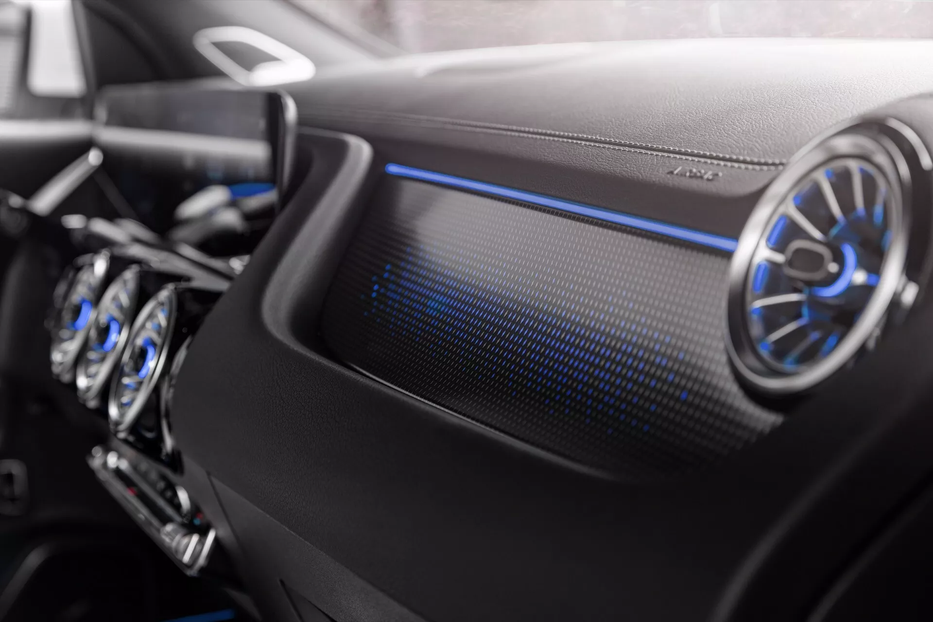 Detailní pohled na interiér Mercedes-Benz EQA 2021 zobrazující palubní desku na straně spolujezdce s kruhovým kovovým větráním a chladným modrým ambientním osvětlením, což zdůrazňuje luxusní design a pokročilé technologické prvky