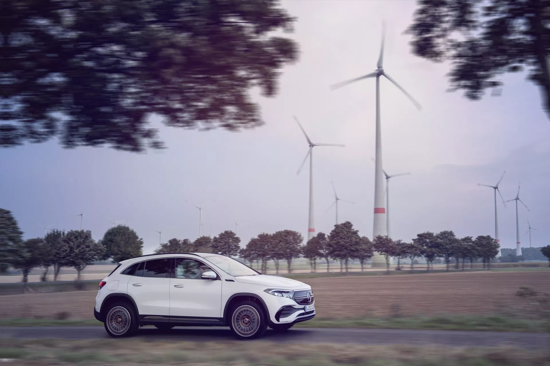 Mercedes-Benz EQA 2021 (243) v pohybu zachycen metodou panoramatické fotografie, představuje jeho moderní SUV profil, složité slitinové kola a ekologicky šetrný design na pozadí větrných turbín