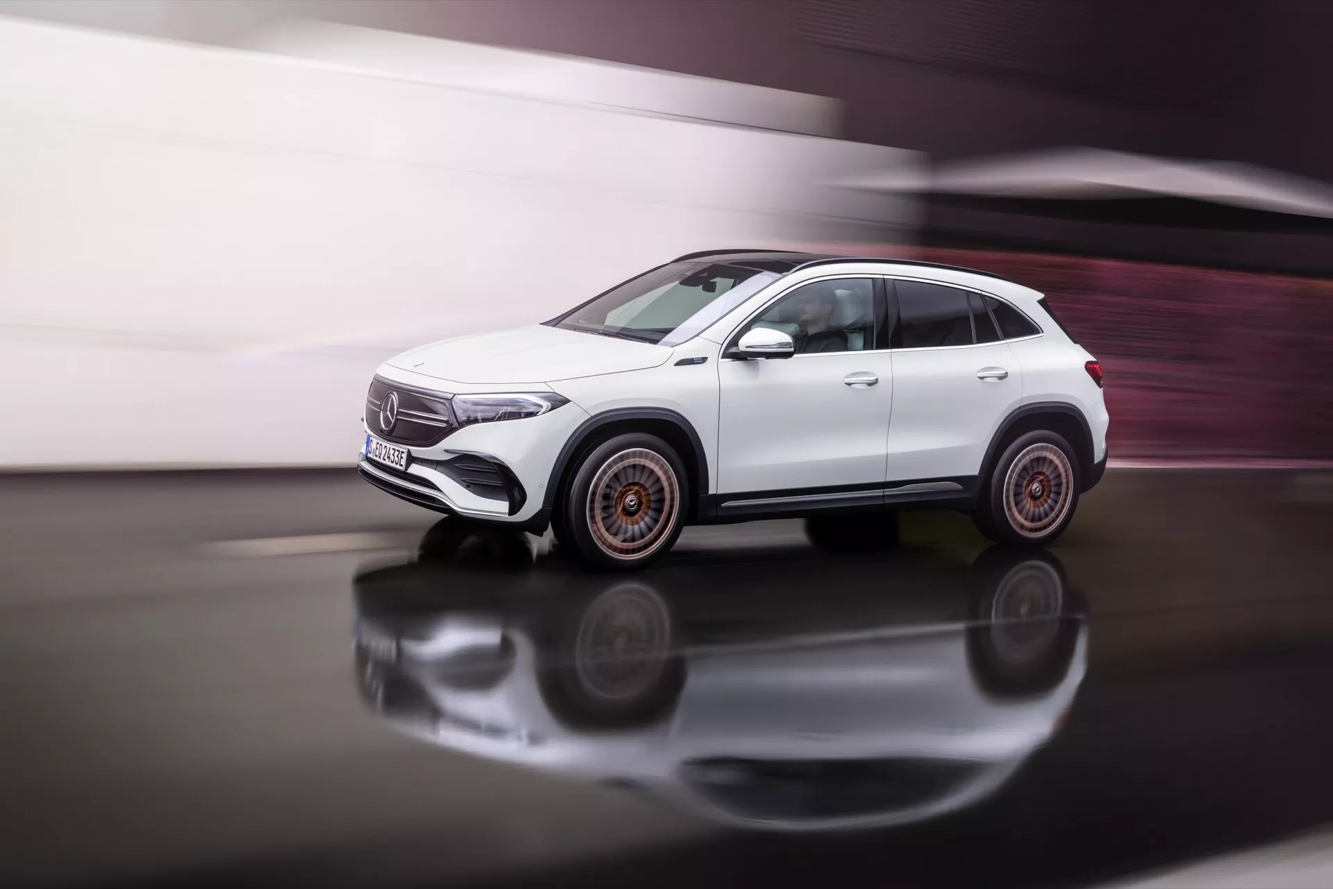 Bílý SUV Mercedes-Benz EQA 2021 v pohybu na odrazivém povrchu s dynamickým bočním pohledem ukazujícím designové linie, tvary oken, dveře a unikátní design kol s jemným rozostřením pohybu a měděnými akcenty