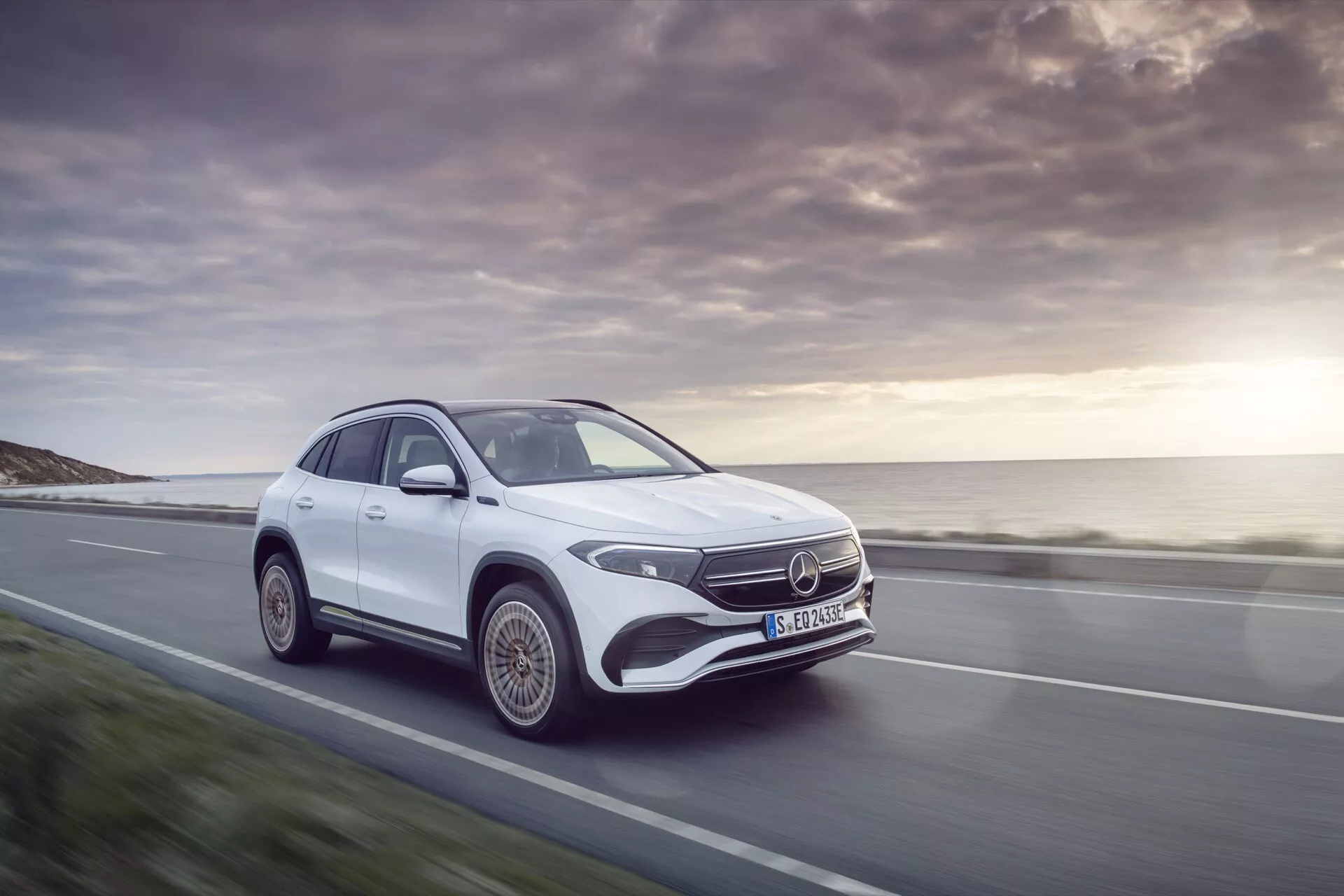 Bílý Mercedes-Benz EQA 2021 ukazující svůj dynamický pohyb a luxusní design na malebné pobřežní silnici, zdůrazňuje jeho LED světlomety, emblém Mercedes a aerodynamický siluetu při západu slunce.