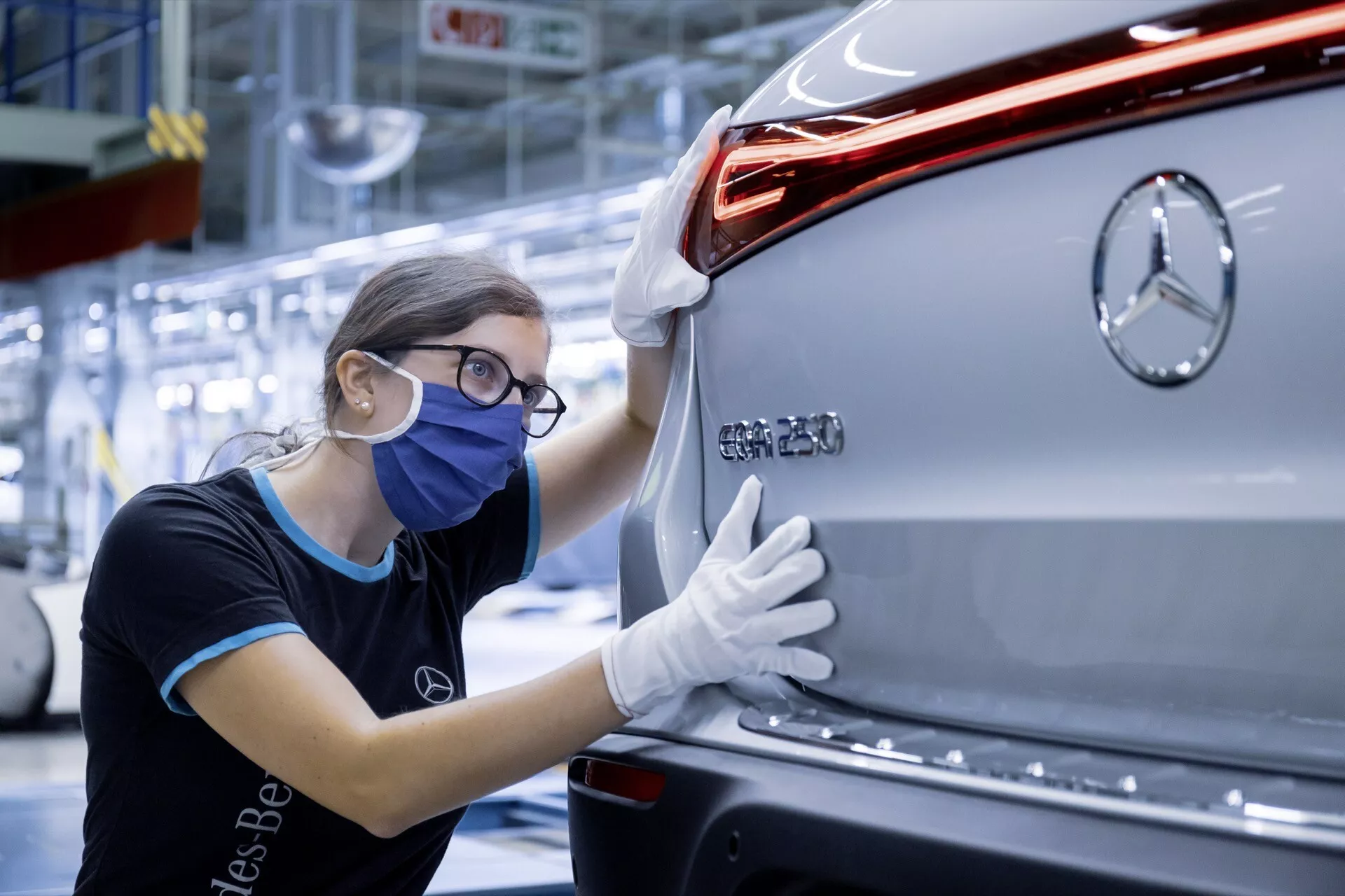 Zadní pohled na stříbrné vozidlo Mercedes-Benz EQC 2021 s výrazným znakem značky, moderním LED zadním světlem, elegantním designem karoserie a zadním nárazníkem, s pracovníkem kontroly kvality provádějícím inspekci vozidla ve výrobním závodě