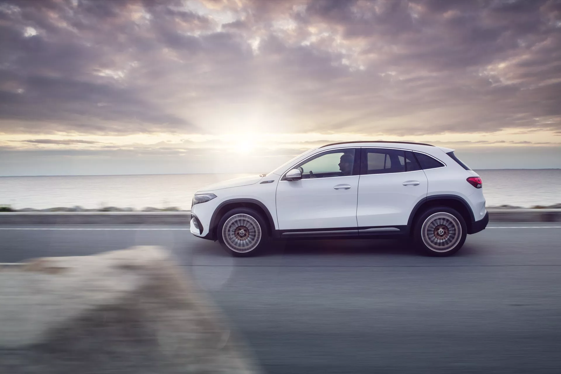 Dynamický boční profil SUV Mercedes-Benz EQA 2021 v bílé barvě, který zvýrazňuje jeho elegantní design a robustní postoj na pobřežní silnici, osvětlen slunečním zářením při západu a technologickými prvkami jako jsou slitinová kola a možné LED osvětlení.