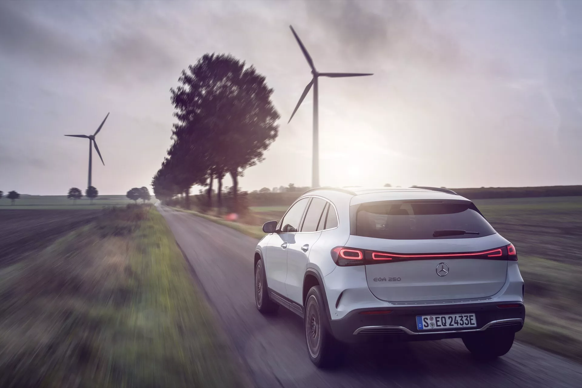 Elektrické SUV Mercedes-Benz EQA 2021 v pohybu, zobrazení ze zadní tříčtvrteční perspektivy zdůrazňující aerodynamický design a moderní technologie, s krajinou větrných turbín využívajících obnovitelnou energii v pozadí