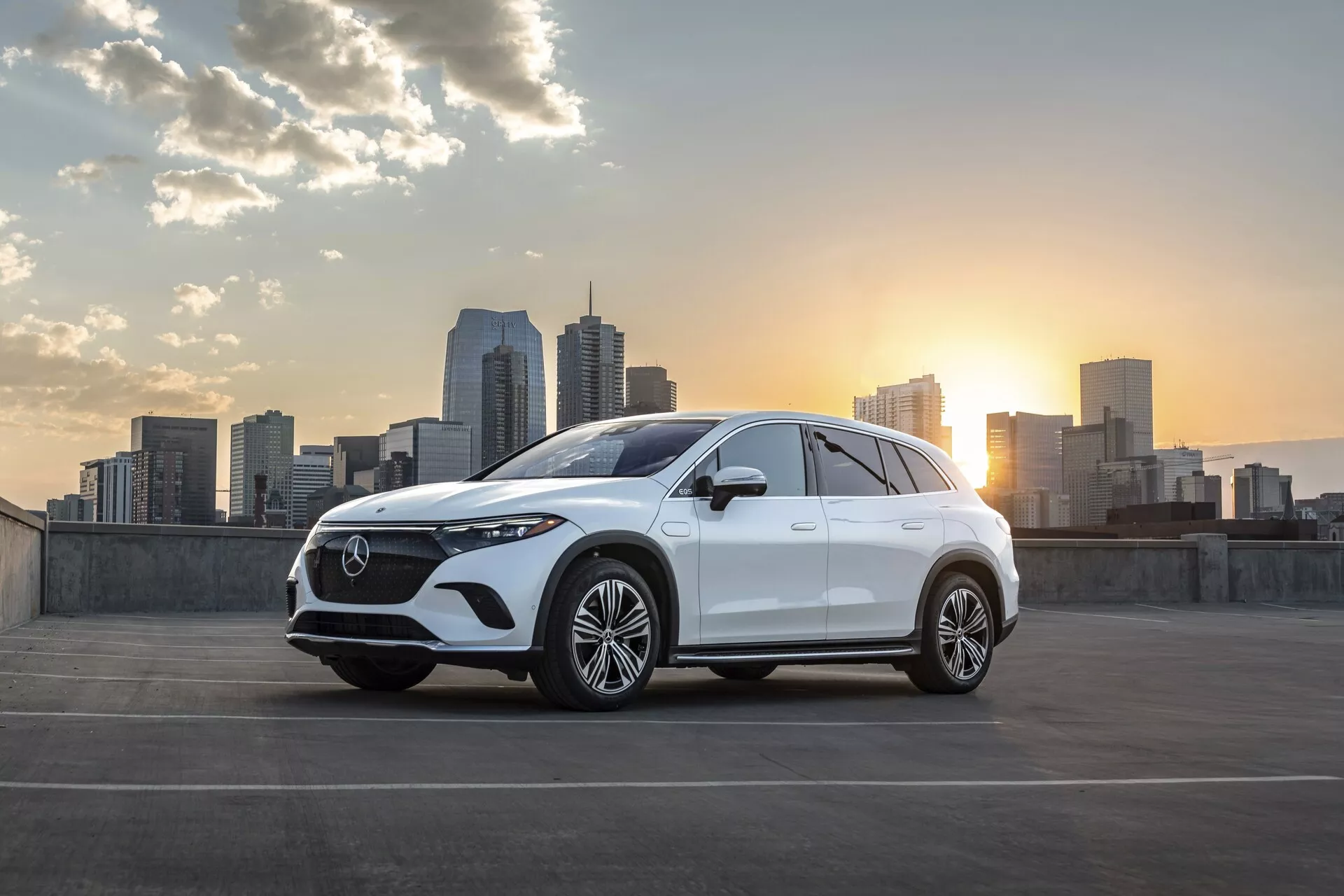 Rafinovaný bílý Mercedes EQS-SUV 2022 zaparkovaný s panoramatem města a zapadajícím sluncem v pozadí. Úhel tříčtvrtečního předního pohledu ukazuje charakteristickou prominentní mřížku s detailním vzorem a centrálním emblémem, elegantní LED světlomety, dvojité přívody vzduchu nebo větrací otvory a velká vícespokeová litá kola. Jemně klesající linie střechy a zvýšená výška světlosti naznačují SUV design. Boční zrcátka pravděpodobně obsahují technologické prvky, jako jsou integrované směrovky nebo kamery pro monitorování mrtvého úhlu. Obrázek je krásně osvětlený měkkými odlesky zapadajícího slunce, které zvýrazňují designové linie vozu. Městské prostředí s mrakodrapy dodává moderní a sofistikovaný nádech, naznačující cílovou skupinu městských zákazníků, kteří oceňují styl a pokročilé technologie.
