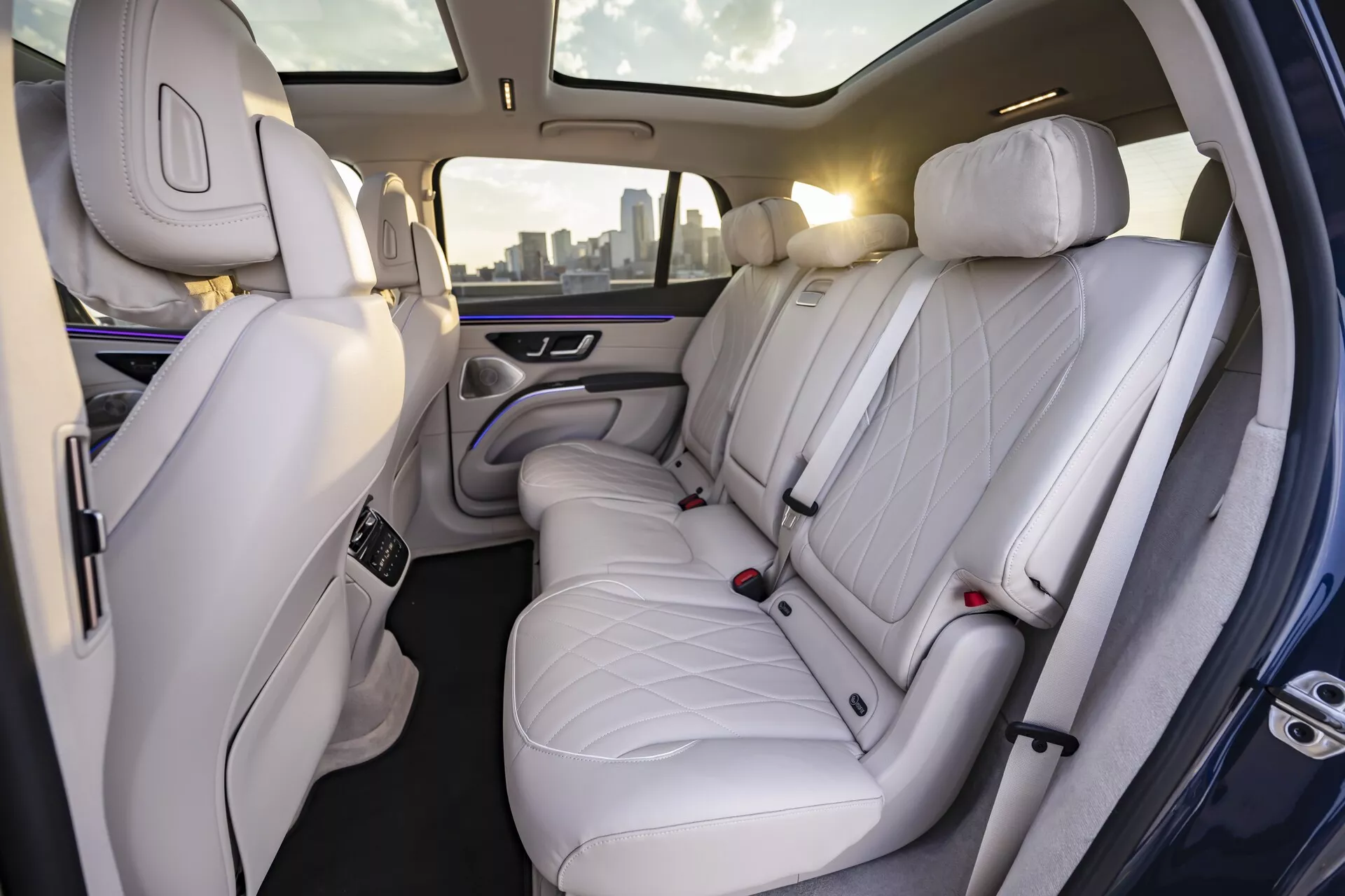 Pohled na interiér Mercedes EQS-SUV 2022, který zobrazuje luxusní zadní prostor pro cestující. Obrázek, pořízený ze strany lehce kloněné doprava, odhaluje vysoce kvalitní světlé nebo světle šedé kožené sedačky s prošíváním ve tvaru diamantů. S třemi hlavovými opěrkami poskytují sedačky dostatečně velký prostor, což svědčí o důrazu na pohodlí a eleganci. Vzadu je vidět i část sedadla spolujezdce, také potaženého prémiovou kůží. Sofistikované ambientní osvětlení s jemným fialovým zářením podél dveřních panelů a centrální opěrky rukou zlepšuje prémiový pocit. Centrální opěrka obsahuje ovládací prvky pravděpodobně pro nastavení klimatu a sedaček a dva držáky na nápoje.