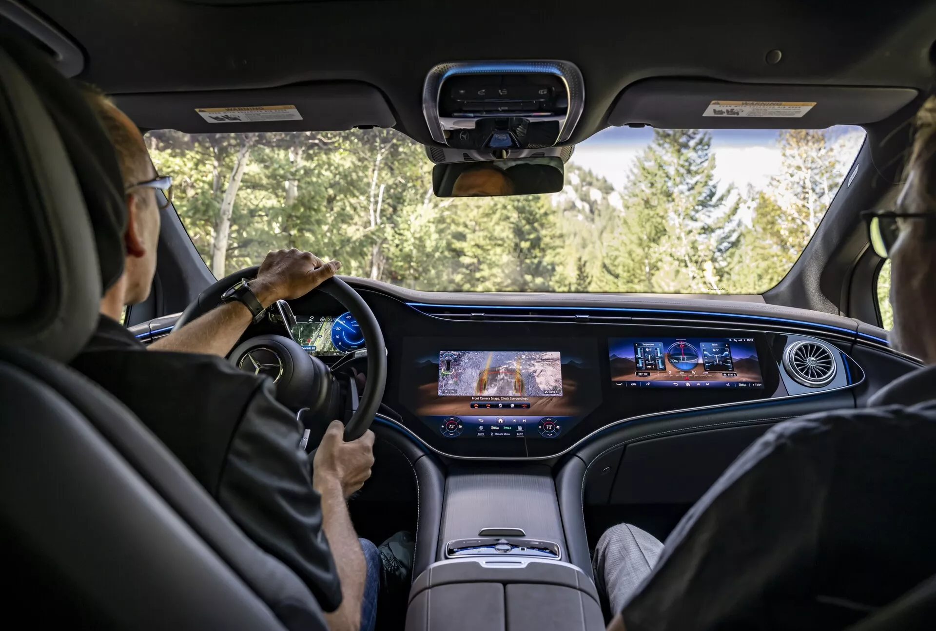 Pohled do interiéru Mercedes EQS-SUV 2022 ze středu zadního sedadla, který zobrazuje moderní palubní desku s nepřerušovaným digitálním displejem, současný volant s montovanými ovládacími prvky, luxusní světle zbarvená kožená sedadla a zvýšenou středovou konzoli. Čelní sklo nabízí výhled na silnici obklopenou lesem, což naznačuje, že auto je v pohybu.