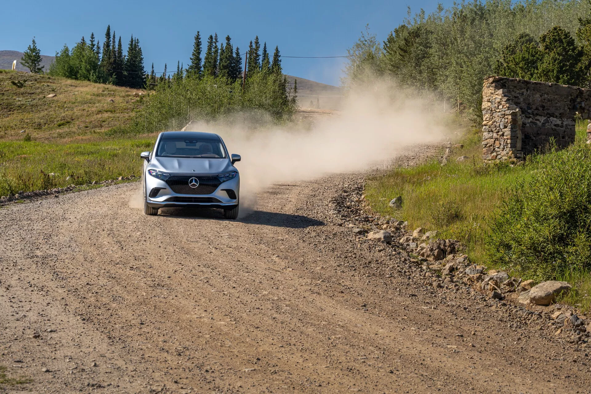 Mercedes EQS SUV 2022 – Moderní hatchback nebo kompaktní SUV jedoucí směrem ke kameře na nezpevněné cestě, zanechávající za sebou oblak prachu. Auto je umístěno uprostřed a zřetelně zobrazí přední mřížku, světlomety, kapotu, čelní sklo a část střechy. Prostředí naznačuje venkovskou nebo nezpevněnou oblast s trávou po obou stranách, jasnou oblohou a pozůstatky staré kamenné struktury v pozadí.