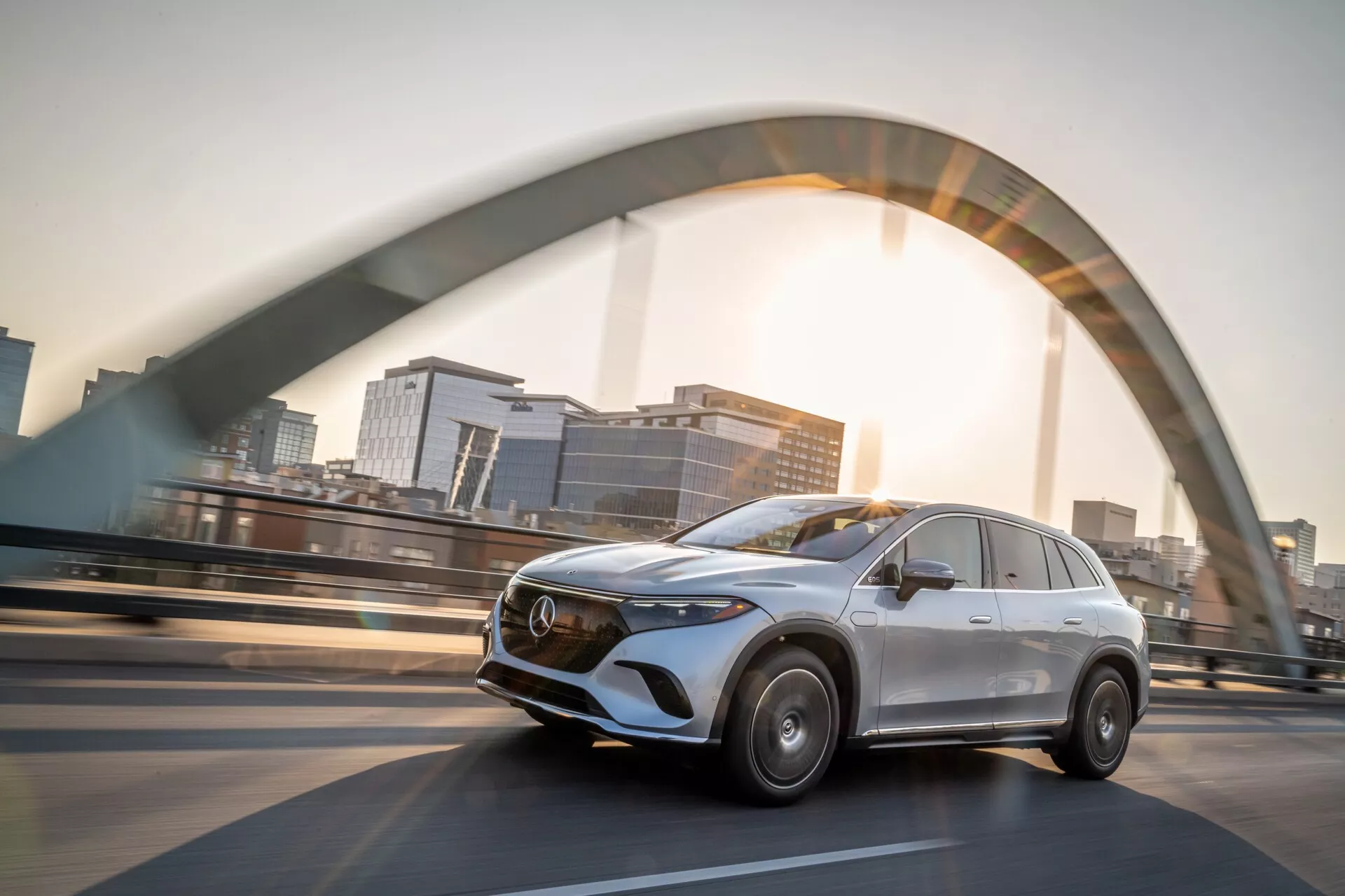 Obrázek ukazuje stříbrný Mercedes EQS-SUV 2022 jedoucí po zvýšeném mostě. Záběr z nízkého úhlu zdůrazňuje moderní design SUV. Na straně řidiče je vidět výrazná chromová mřížka s emblémem, elegantní linie, stylizované LED světlomety a rozostření pohybu kol, což naznačuje rychlost. Slunční záře a slabý obrys města přidávají dramatický efekt, podtrhujícej luxus a pokročilé inženýrství.