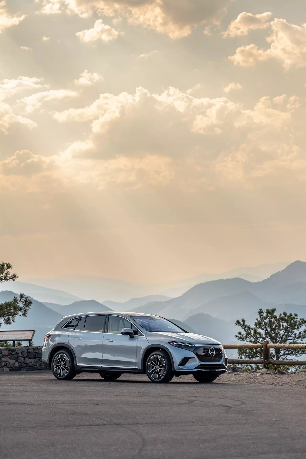 Moderní Mercedes EQS-SUV 2022 zaparkovaný na malebném vyhlídkovém místě, zvýrazňující jeho pravý boční profil s hliníkovými koly, klikami dveří, postranními zrcátky, okenními lištami a koly lehce natočenými doleva. Fotografováno za úsvitu nebo soumraku s teplým, rozptýleným osvětlením, na pozadí panoramatických hor a lehce zatažené oblohy, zdůrazňující luxus, dobrodružství a vysoce kvalitní řemeslné zpracování.
