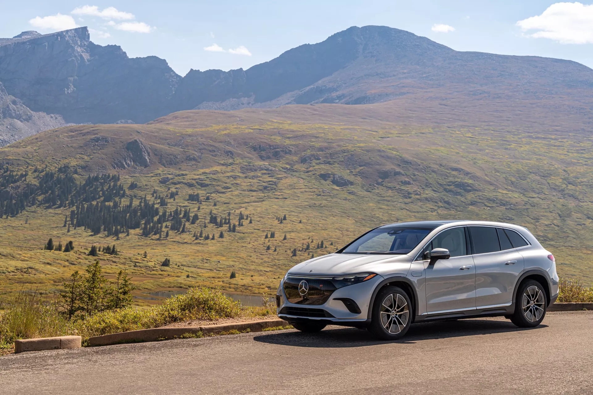 Obrázek zobrazuje moderní Mercedes EQS-SUV 2022 umístěný na asfaltové silnici s malebnou horskou scenérií na pozadí pod jasnou oblohou se rozptýlenými mraky. Pohled z tří čtvrtin zepředu odhaluje klíčové prvky, jako je mřížka chladiče s emblémem, pokročilá sestava světlometů a design předního nárazníku s čidly nebo kamerami. Aerodynamické linie na kapotě a bocích spolu s bočním profilem zobrazujícím litá kola, boční zrcátka, charakterové linie a sklon střechy zdůrazňují design a technologické prvky vozidla.