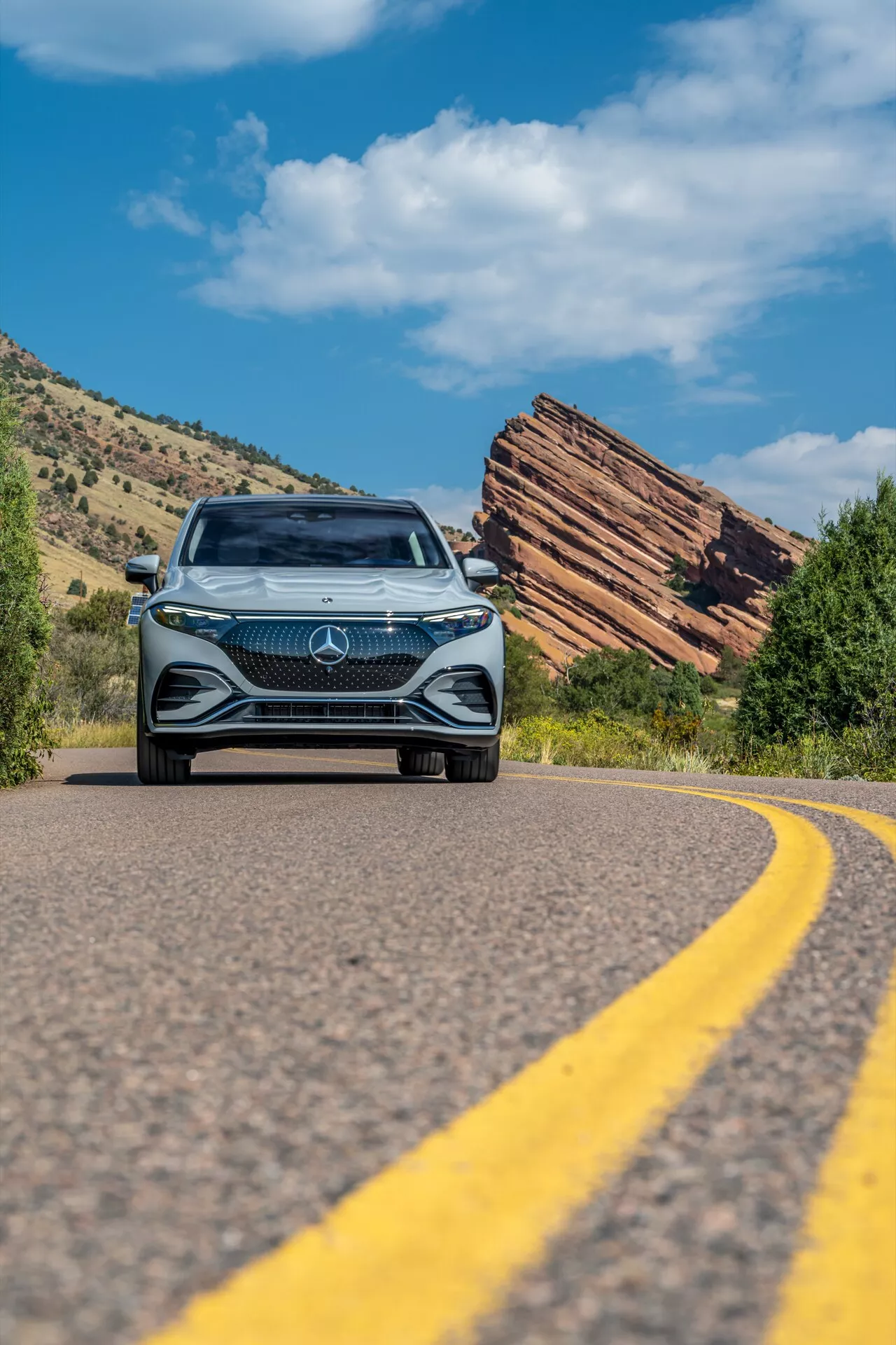 Nízký úhel záběru Mercedes EQS SUV 2022 přibližující se na zatáčející silnici, ukazující odvážnou přední mřížku s velkým hvězdným emblémem, složitý diamantový design, LED světlomety a denní svícení, zasazené proti jasně modré obloze a drsnému přírodnímu terénu se stratifikovanými skalními útvary.