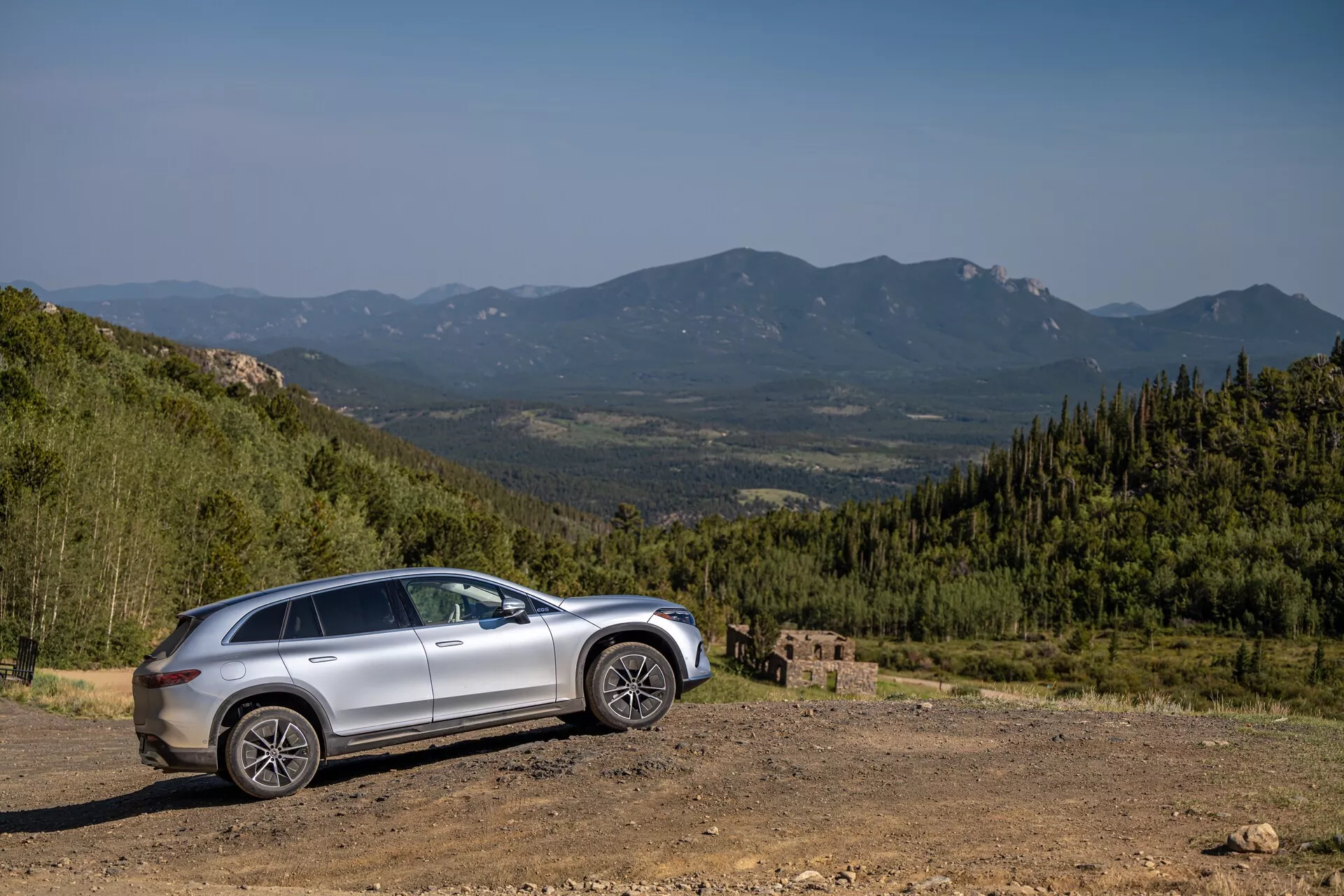 Boční profil stříbrného Mercedesu EQS-SUV 2022 na nerovném terénu s malebným horským pozadím, vyfocený pod úhlem 30 až 40 stupňů. Vlastnosti zahrnují vysokou světlou výšku, zvýšenou jízdní výšku, velká robustní kola, tvarované linie na dveřích, pravděpodobně střešní okno, zrcátka s integrovanými směrovkami, tónovaná okna a moderní LED světlomety a zadní světla.