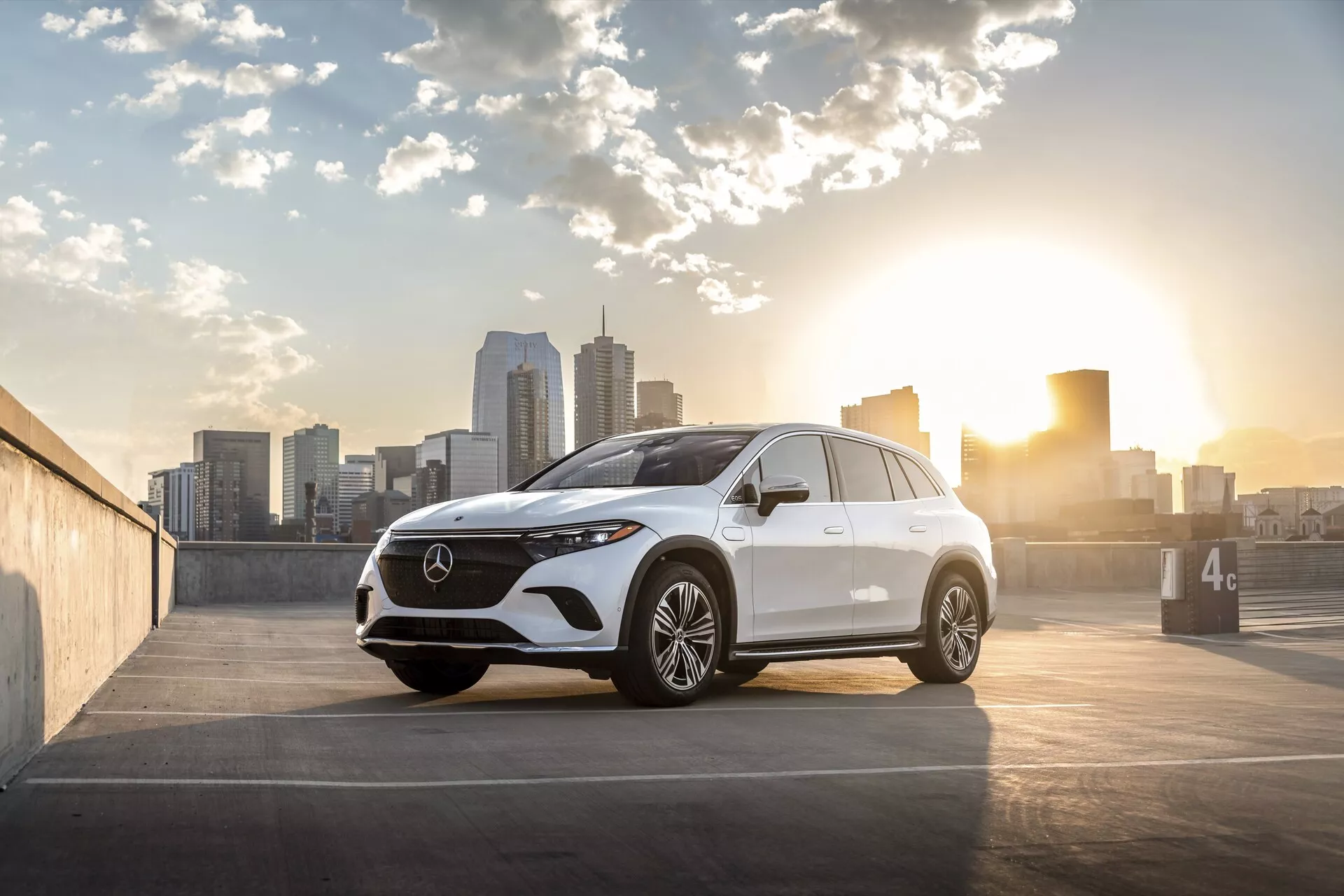 Obrázek ukazuje moderní Mercedes EQS-SUV 2022 umístěný na venkovním parkovišti s panoramatem města v pozadí, zachycený buď při východu nebo západu slunce s teplým osvětlením zezadu. Pohled tří čtvrtin zepředu zdůrazňuje masku chladiče, světlomety, design předního nárazníku, kapotu a stranu řidiče, focený z nízkého úhlu blízko úrovně kol. Detaily zahrnují prominentní mřížku s velkým emblémem, elegantní LED světlomety, přední kameru nebo snímačový klastr, vícepaprsková alu kola, integrované směrovky na bočních zrcátkách, jemné konturové linie pro aerodynamiku a mírně klesající linii střechy. Složení fotografie umisťuje auto na pravou stranu s panoramatem města a sluncem vlevo, přidávající odlesk a uměleckou přitažlivost, vhodné pro kontexty automobilismu, luxusu a městského životního stylu.