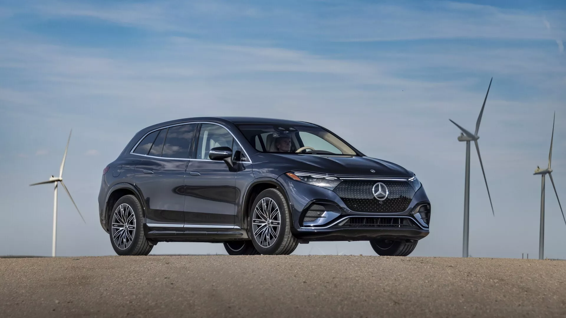 Boční profil SUV Mercedes EQS 2022 v tmavé barvě, zaparkovaného na rovné ploše. Obrázek, pořízený z mírného úhlu přední čtvrtiny, ukazuje elegantní a moderní design se zakřivenými liniemi a hladkými povrchy, které zdůrazňují aerodynamickou účinnost. Významné prvky zahrnují výraznou masku s centrálním logem značky, elegantní LED světlomety, nasávací otvory nebo senzory v spodním nárazníku, sofistikovaná vícepaprsková slitá kola, boční zrcátka s integrovanými směrovkami, chromově lemovaná okna a jemně se svažující střešní linii s nosiči na střeše. V pozadí jsou větrné turbíny, které naznačují ekologickou nebo elektrifikovanou povahu vozu.