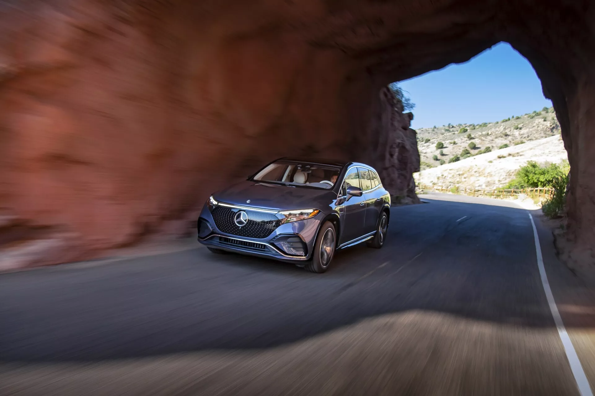 Obrázek vozu Mercedes EQS-SUV 2022 vyjíždějícího z tunelu na silnici, zachycený z nízkého úhlu se zaměřením na přední levý bok, zdůrazňující moderní design s výraznou maskou, logem, denními LED světly, vícepaprskovými slitinovými ráfky a kovovým lakem. Vůz je v pohybu, což naznačují rozmazané pozadí a kola. Okolí zobrazuje skalnatý terén s řídkou vegetací, což naznačuje horskou nebo pouštní oblast za jasného počasí.