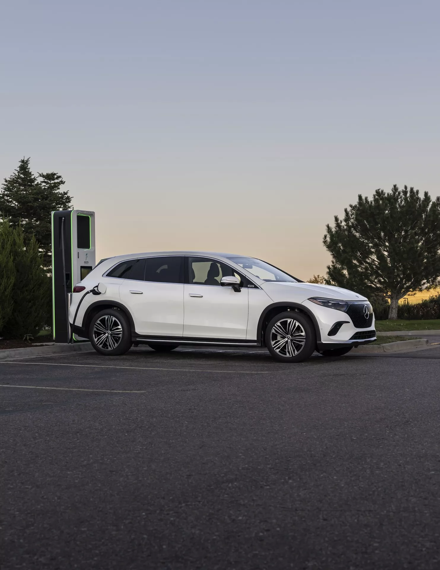 Moderní Mercedes EQS-SUV 2022 u nabíjecí stanice za soumraku, fotografovaný z bočně-předního úhlu, zdůrazňující jeho elegantní profil, slitinková kola a nabíjecí port.