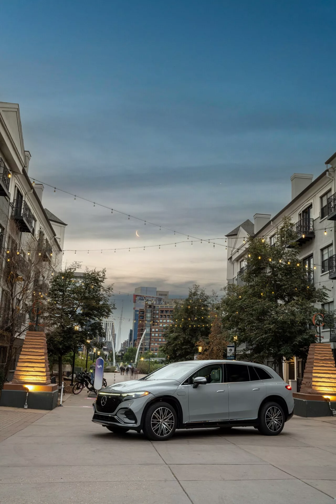 Přední tří čtvrtinový pohled na stříbrný Mercedes EQS-SUV 2022 s nápadnou mřížkou s emblémem, LED denními světly, litými koly a karoserií. Auto je zaparkováno v luxusním městském prostředí s dekorativním osvětlením a budovami, což zvýrazňuje jeho luxusní a moderní design.