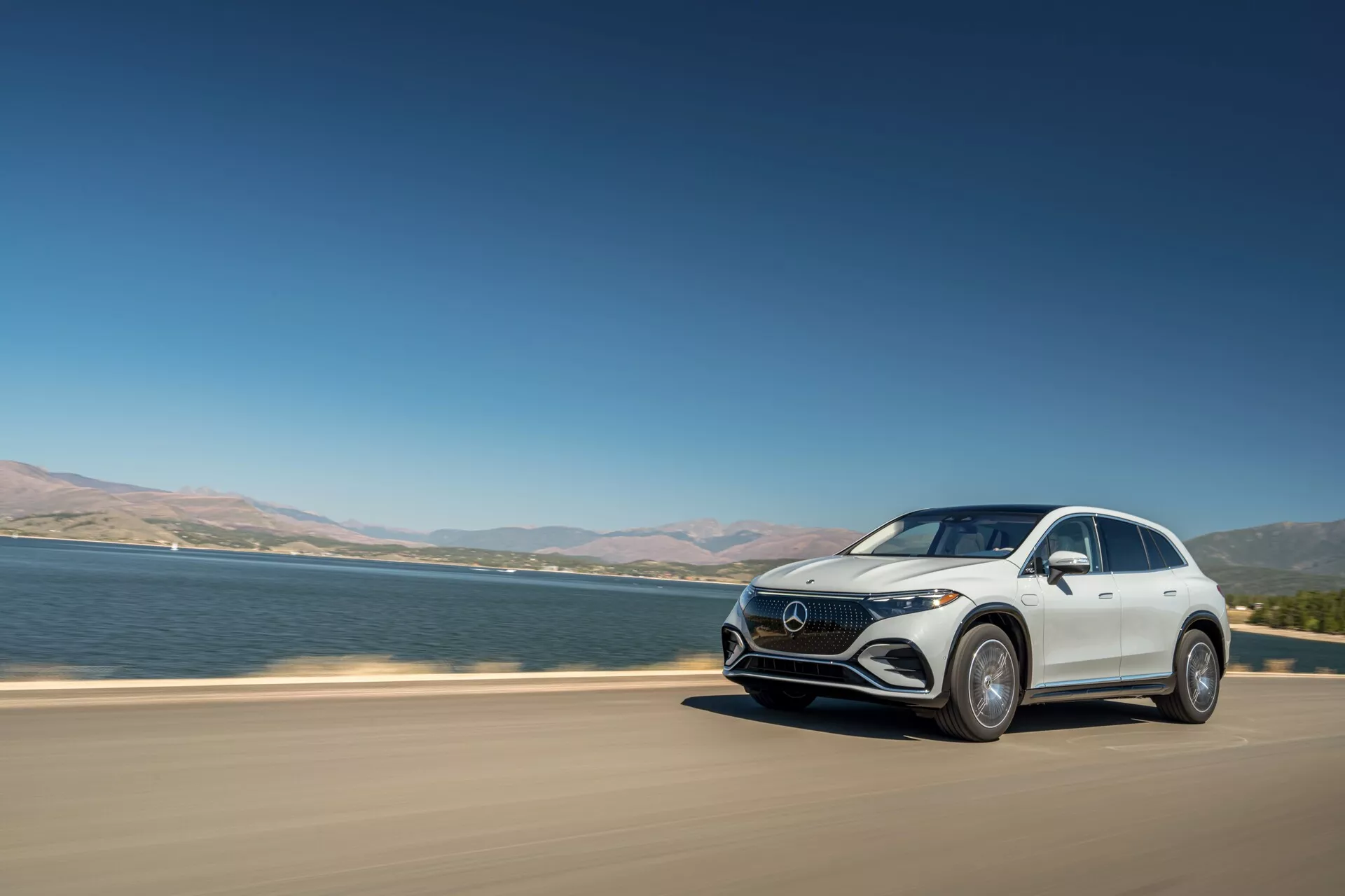 Moderní luxusní crossover SUV Mercedes-Benz EQS jedoucí po asfaltové silnici poblíž velkého vodního tělesa. Obraz zachycuje vůz z tříčtvrtečního čelního pohledu za jasného denního světla a zdůrazňuje funkce jako LED světlomety, charakteristickou masku, aerodynamický design a velká litá kola. Vozidlo je v pohybu, což naznačuje dynamiku, s jezerem nebo řekou v pozadí a jasnou oblohou nad hlavou, což je charakteristické pro prémiové elektrické modely.