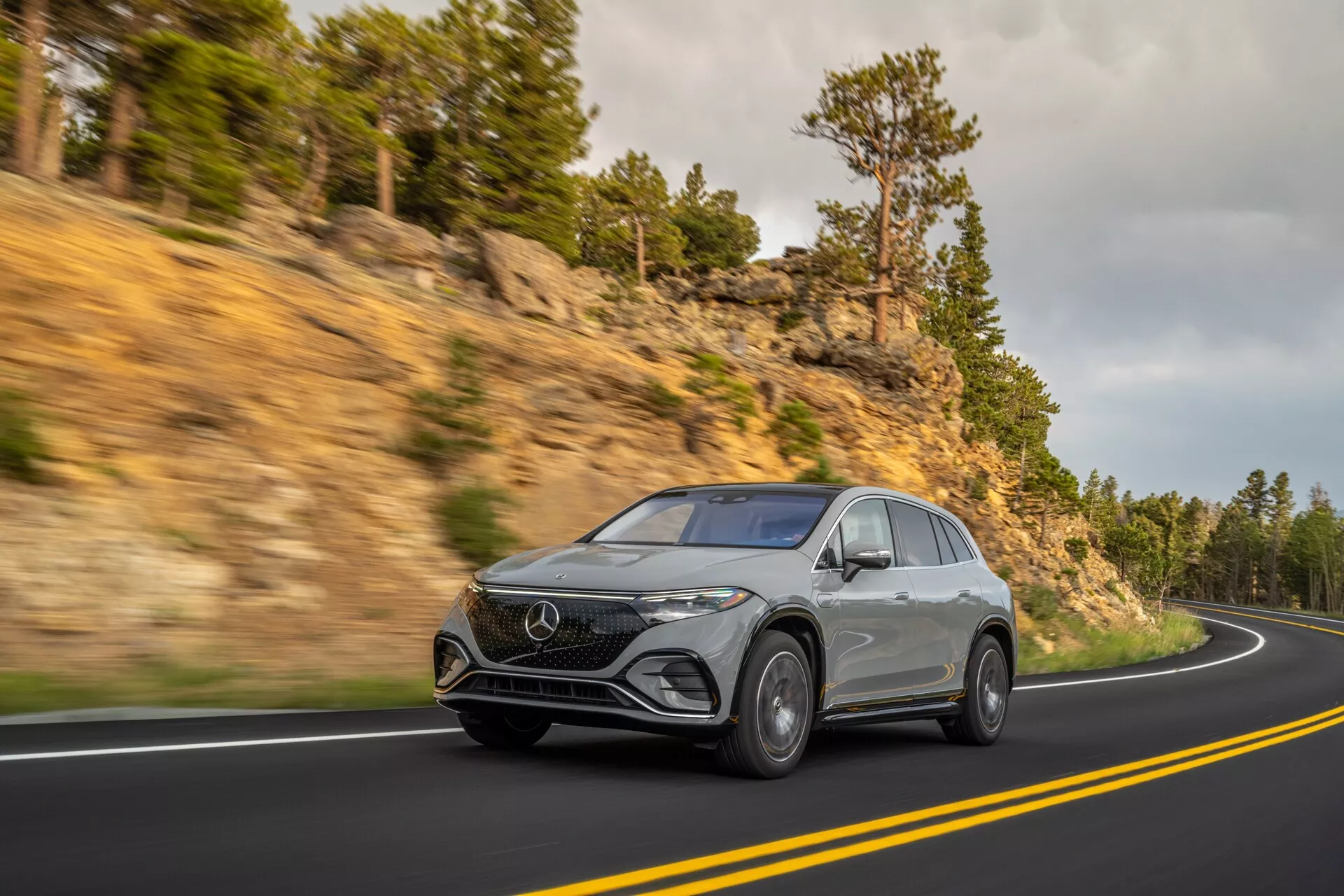 Dynamický snímek stříbrného Mercedesu EQS SUV 2022 na klikaté horské silnici, s pohledem z tříčtvrtečního předního úhlu, aby vynikla jeho výška a přítomnost na silnici. S moderními LED světlomety, výrazným předním znakem, luxusními slitinovými koly a aerodynamickými liniemi potvrzujícími současný design.