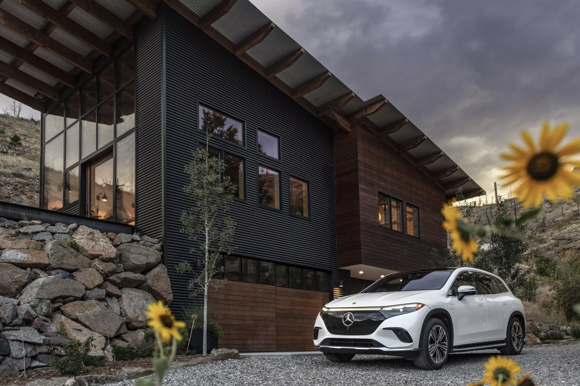 Obrázek luxusního SUV Mercedes-Benz EQS 2022 zaparkovaného před moderním domem s oblačným nebem v pozadí. Záběr z nízkého úhlu zdůrazňuje postoj a velikost vozidla, zaměřuje se na přední masku, sestavu světlometů, přední nárazník, kontury kapoty a levou stranu řidiče až po zadní dveře. Mezi prvky patří LED denní svícení a elegantní kola, což přispívá k pocitu luxusu a pokročilé technologie. Bílé SUV kontrastuje s tmavým domem a skalnatou krajinou, obklopené mírně rozmazanými květinami díky malé hloubce ostrosti, která zvýrazňuje SUV jako hlavní bod.