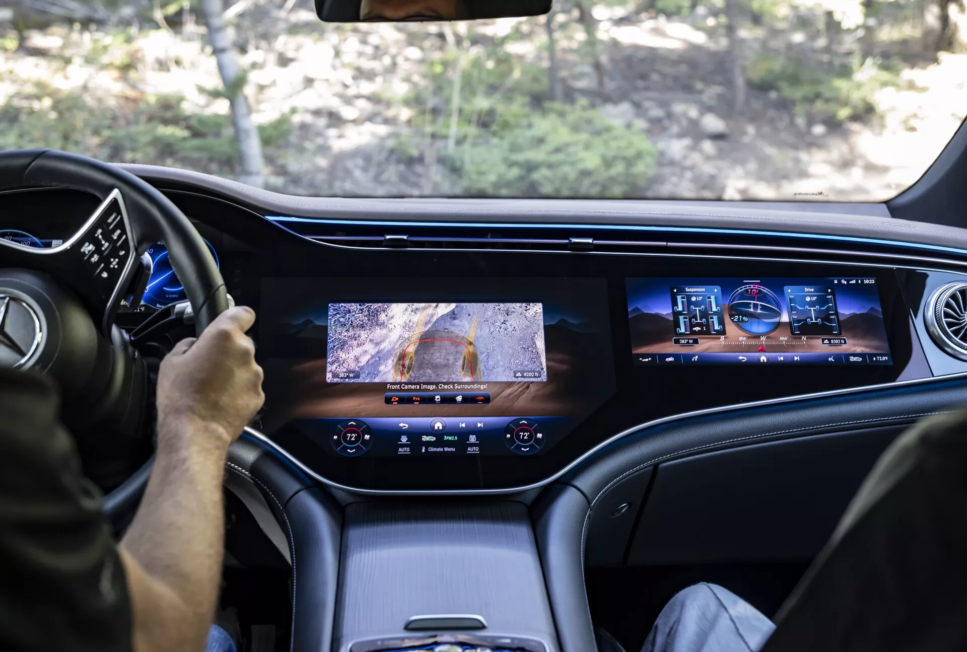 Pohled ze zadního sedadla Mercedesu EQS-SUV 2022 ukazuje přední interiér; zobrazuje řidiče používajícího multifunkční volant, digitální přístrojovou desku, velkou centrální obrazovku s kamerovým zobrazením, sekundární infotainmentovou obrazovku pro spolujezdce, prémiové kožené nebo kvalitní materiály kabiny a moderní design větracích otvorů, zdůrazňující luxusní a pokročilé technologické prvky vozu.