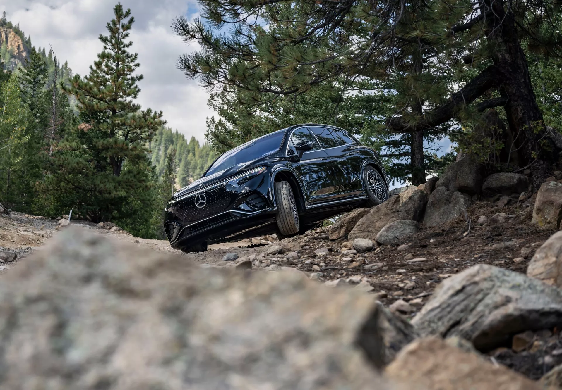 Nízký záběr černého SUV Mercedes-Benz EQS 2022 v drsné venkovní scéně, ukazující jeho levý předek s částečným pohledem na pravou stranu. Obrázek zvýrazňuje výrazné světlomety, mřížku s Mercedes emblémem, litá kola a robustní pneumatiky na šterkovém cestě, zdůrazňující schopnost v terénu a luxusní design.