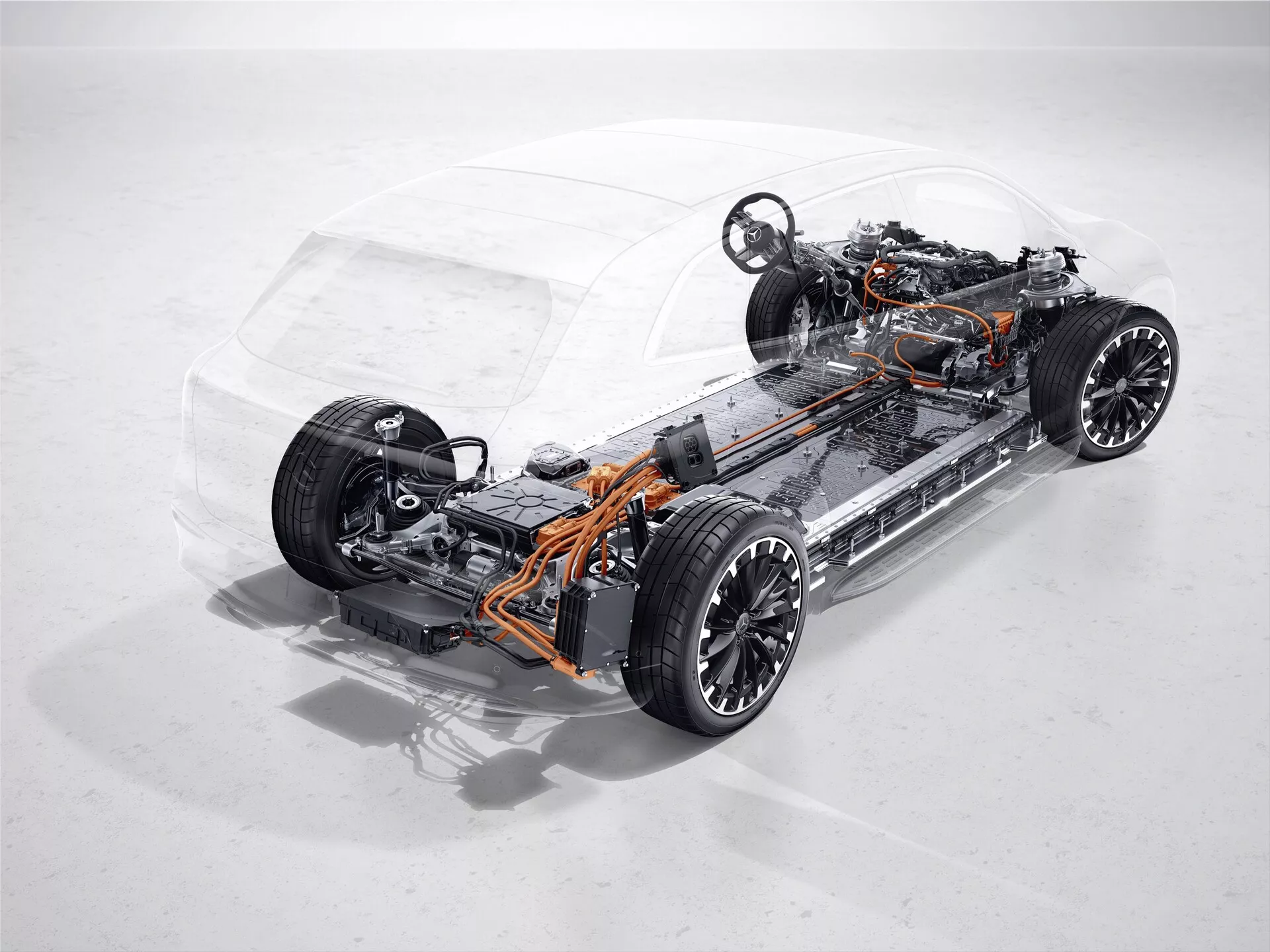 Studiová fotografie průhledného podvozku a pohonu modelu Mercedes EQS SUV 2022. Obrázek je pořízen z mírného nadhledu, zvýrazňující přední levobok. Fotografie ukazuje vnitřní strukturu, včetně předních a zadních náprav, odpružení, kol a nízko umístěného bateriového bloku typického pro moderní elektrická vozidla. Viditelné oranžové kabely naznačují vysokonapěťový systém. Pohonná jednotka vozu, charakterizovaná absencí tradičního spalovacího motoru, naznačuje přítomnost jednoho nebo více elektromotorů. Absence výfukového systému a palivové nádrže potvrzuje elektrickou povahu vozidla. Nezávislé odpružení naznačuje pokročilé jízdní a jízdní vlastnosti.