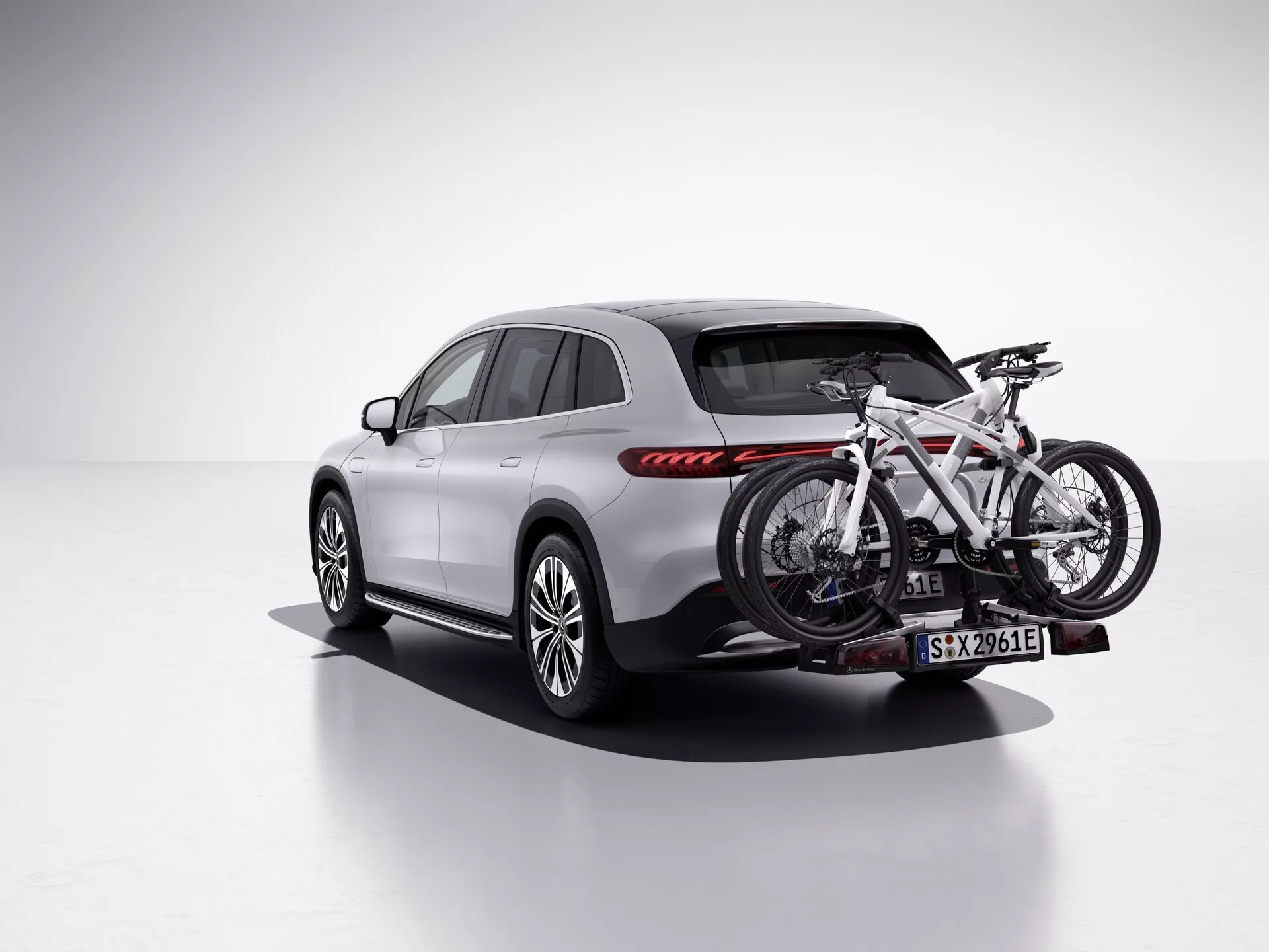 Obrázek zachycuje SUV Mercedes EQS z roku 2022 ve studiovém prostředí s čistým, bílým pozadím, pohledem z zadního tříčtvrtečního úhlu ukazujícím stranu pro cestující. Vozidlo je vybaveno nosičem kol na zadním tažném zařízení, který drží jedno kolo. SUV je elegantního, moderního designu s hladkými liniemi, výrazným zadním světelným klastrem s horizontálními červenými LED brzdovými světly, vícepaprskovými litými koly a karoserií lakovanou v světlé barvě, možná stříbrné nebo šedé, kontrastující s černým spodním ozdobným dílem a obložením kol. Boční zrcátka, střešní ližiny a okenní lišty jsou v tmavším tónu, s panoramatickým střešním oknem dodávajícím prémiový pocit. Tento obrázek zdůrazňuje funkce jako LED osvětlení, sportovní design kol a nosič kol, což ho činí vhodným pro kategorie jako Vozidla, Automobilová technologie, Venkovní/sportovní vybavení a Luxusní SUV.