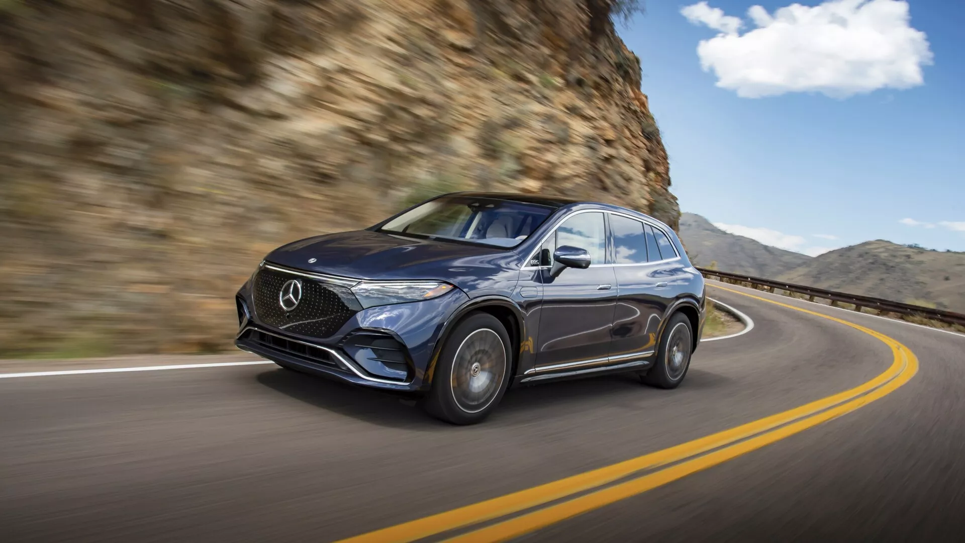 Obrázek tmavého Mercedesu EQS SUV 2022 v pohybu, zachycený z předního levého tříčtvrtečního pohledu. Auto jede po zakřivené silnici s skalní stěnou na pravé straně. Důraz je kladen na luxusní design s výraznou mřížkou s emblémem Mercedes, elegantními světlomety, lesklým povrchem a částečně viditelnými litými koly. Rozmazané pozadí zvyšuje pocit pohybu, podtrhující jízdní schopnosti a prémiový vzhled vozu v přirozeném jízdním prostředí.