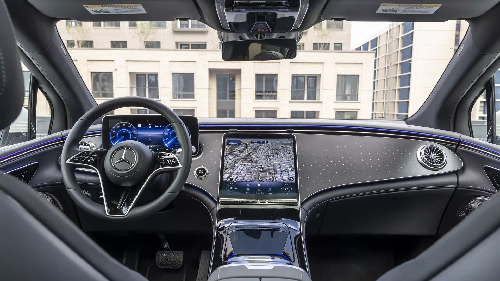 Interiérový pohled na Mercedes EQS SUV 2022 z pohledu z zadního sedadla, zachycující přístrojovou desku, multifunkční volant s plochým spodkem a kovovými akcenty a středovou konzolu s prodlouženým dotykovým infotainment systémem. Digitální přístrojový panel je osvětlený a minimalistický elektronický volič převodových stupňů, úložné prostory a držáky nápojů jsou viditelné. Luxusní interiér vozu zahrnuje materiály vysoké kvality, leštěné dřevěné obložení a částečně viditelnou panoramatickou střechu, ukazující moderní automobilový design a pokročilé technologické funkce.