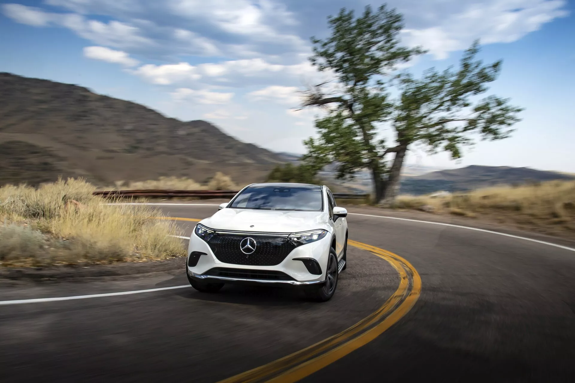 Dynamický pohled na Mercedes EQS-SUV 2022 z předního tříčtvrtečního úhlu v pohybu, který ukazuje design mřížky, světlometů, kapoty, kola a bočních kontur. Auto zabírá pravou stranu rámu, pohybuje se doleva s rozmazaným pozadím naznačujícím pohyb. Viditelné funkce zahrnují LED denní svícení, design světlometů, nárazník s integrovanými přívody vzduchu nebo senzory a aerodynamicky tvarovaná boční zrcátka. Silnice a přírodní krajina naznačují použití na dálnici nebo pro turistiku, čímž se zvýrazňuje výkon, luxus a potenciálně pokročilá technologie vozidla.
