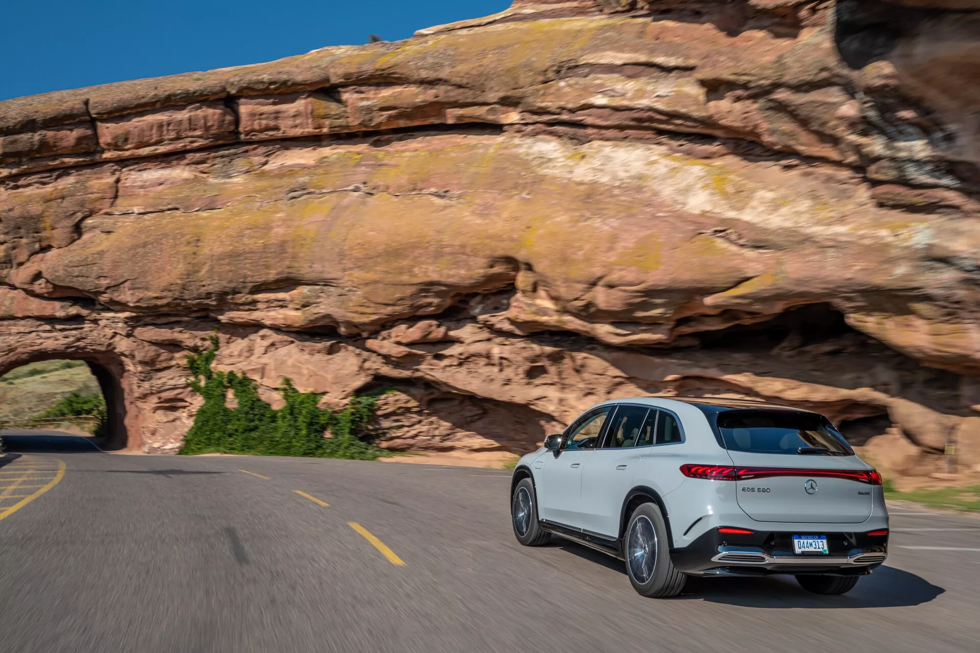 Bílý Mercedes EQS-SUV 2022 jedoucí na zakřivené dvouproudé silnici, fotografovaný ze zadního tříčtvrtečního pohledu, ukazujícího zadní světla a profil spolujezdce. Vlastnosti zahrnují výrazný zadní spoiler, dvoujité výfukové potrubí, stříbrné ozdobné lišty, chromované akcenty a vícepaprsková slitinová kola. Rozmazání pohybu na kolech a pozadí zachycuje dynamickou jízdu SUV v scénické horské oblasti pod jasnou modrou oblohou.