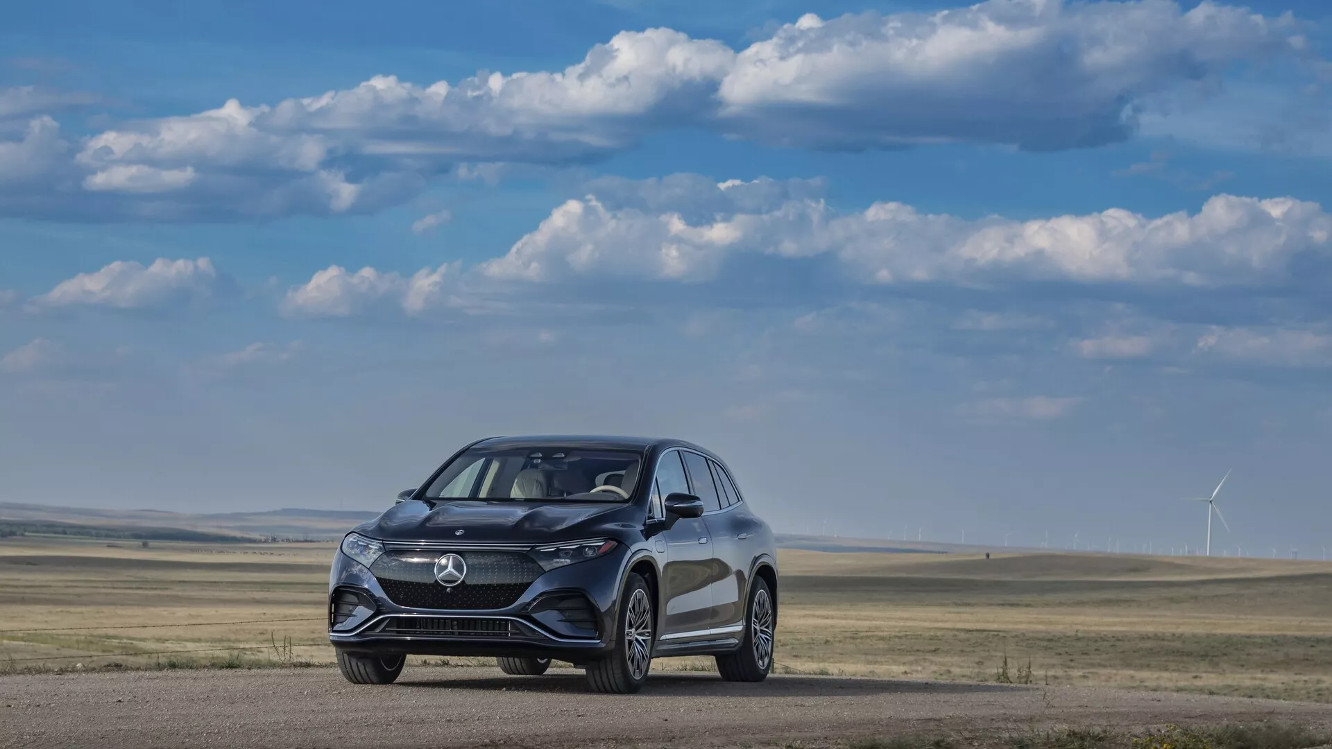 Obrázek zobrazuje Mercedes EQS SUV 2022 z předního tříčtvrtečního úhlu, umístěný na rovném povrchu s trávníkem a větrnými turbínami v pozadí pod jasnou, částečně oblačnou oblohou. Elegantní, aerodynamicky navržené vozidlo vyniká moderní mřížkou, LED světlomety a koly zaměřenými na výkon. Viditelné detaily naznačují pokročilé asistenční systémy řidiče včetně senzorů a kamer integrovaných do předního nárazníku a bočních zrcátek.
