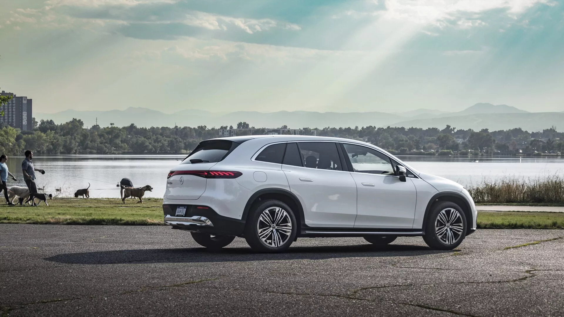 Profilový pohled na luxusní Mercedes EQS SUV z roku 2022 fotografovaný na asfaltové silnici. SUV má velká vícepaprsková slitá kola, elegantní linku oken zužující se směrem dozadu s tónovanými okny, výrazné aerodynamické linie karoserie, pokročilá boční zrcátka, svažující se střechu s nosičem a obtočené LED zadní světlomety. Vozidlo je ponořeno do teplého slunečního svitu, což zvýrazňuje jeho moderní design na pozadí s tělesem vody, stromy a procházejícími lidmi.