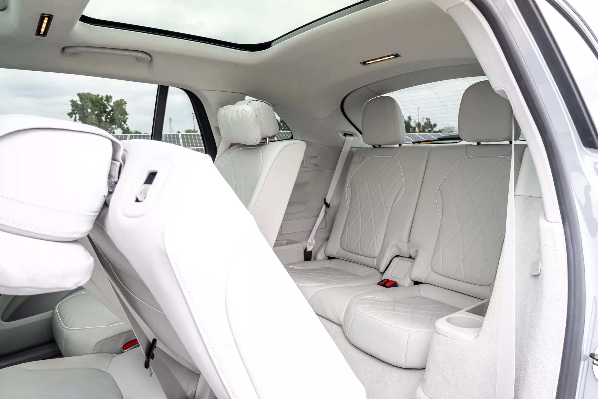 Interiérový pohled na luxusní Mercedes EQS-SUV 2022, zaměřující se na zadní prostor pro pasažéry s bílými perforovanými koženými sedadly, panoramatickým střešním oknem, dveřmi s odpovídající kůží a mřížkami reproduktorů, zadními ventilačními otvory a stropními světly. Otevřené dveře odhalují prostorné a pohodlné uspořádání sedadel pro tři pasažéry.