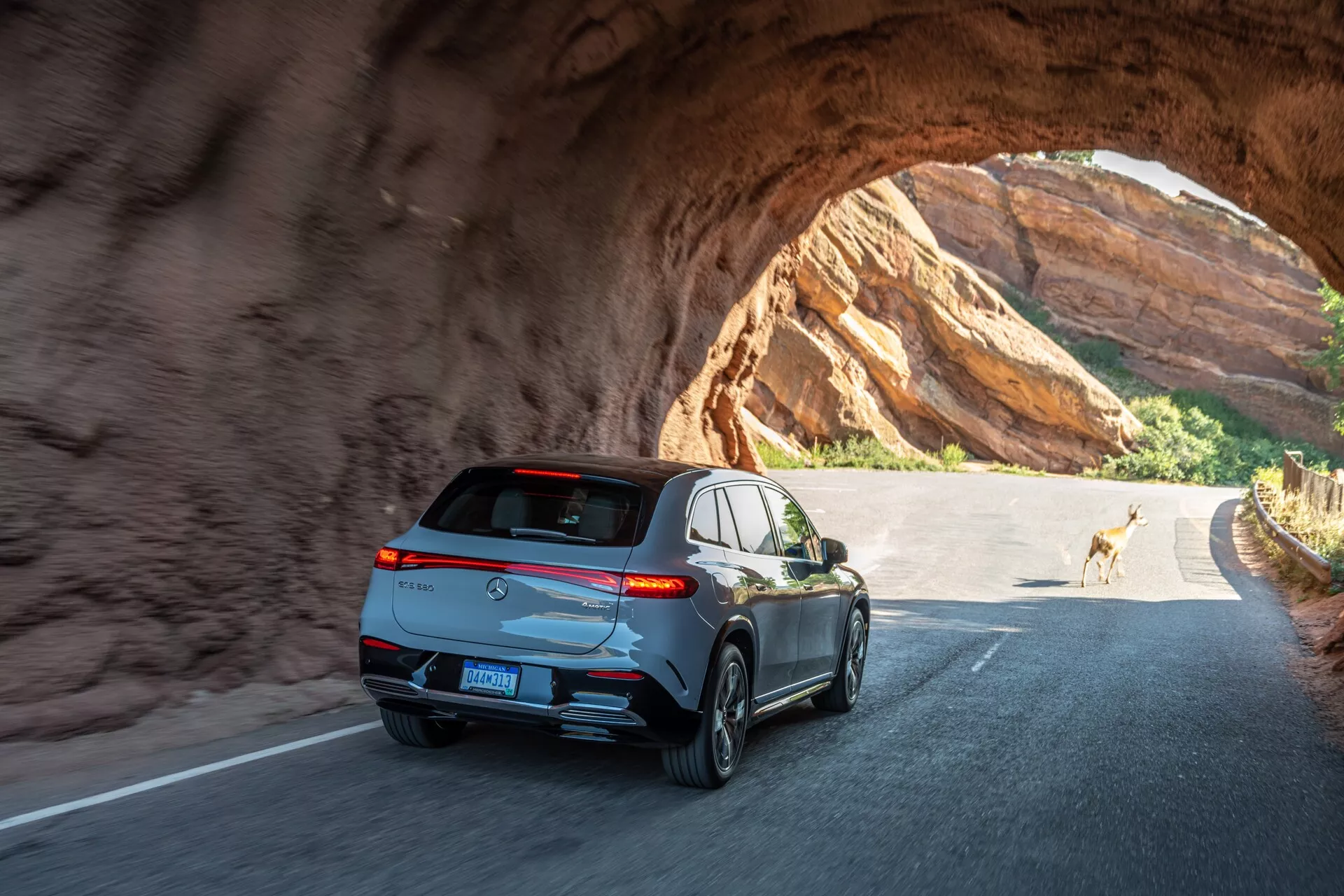 Zadní a úhlový pohled na elegantní, moderní SUV projíždějící skálovým tunelem za slunečného dne. Klíčové prvky zahrnují horizontální obkroužené zadní světlomety, sportovní spoiler, žraločí anténu, reflexní lemování oken, dvojité výfukové trubky, pětiramenná litá kola a centrálně umístěnou poznávací značku pod tónovaným zadním oknem. Vozidlo vykazuje pokročilou světelnou technologii a pravděpodobně je vybaveno funkcemi jako je zadní kamera a parkovací senzory. Fotografie zachycuje pohyb a pocit venkovního dobrodružství se srnkou v pozadí.