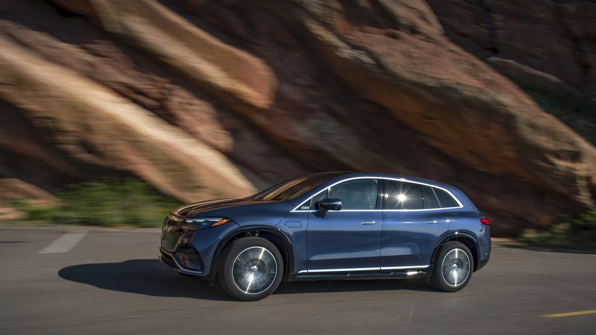 Mercedes EQS-SUV 2022 v pohybu, boční pohled. Jízda zleva doprava po dlážděné cestě s pozadím skalní stěny, naznačující hornatý terén. Pravý boční pohled ukazuje moderní design: viditelné přední a zadní dveře pro cestující, dobře definovaná boční okna s obložením, slitá kola a výrazné boční zrcátko. Kovová barva, pravděpodobně LED nebo HID světlomety, zapuštěné kliky dveří. Možné pokročilé technologické prvky: bezpečnostní systémy, infotainment, elektrický/hybridní motor. Mírné rozmazání pohybu ukazuje dynamický výkon, denní snímek s přirozeným osvětlením.