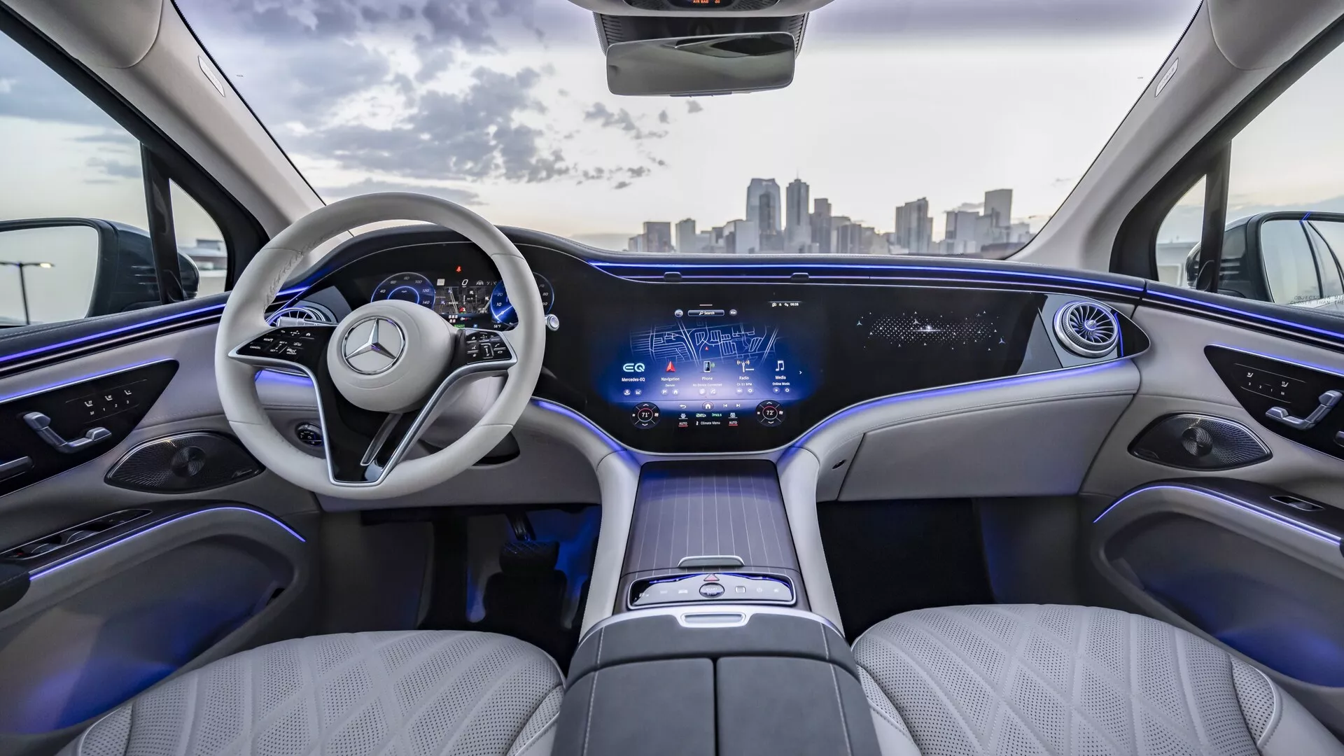 Pohled na interiér Mercedesu EQS SUV 2022 zezadu, s luxusním světlým koženým čalouněním, elegantní palubní deskou s kontinuálním digitálním displejem, moderním dvouramenným volantem s emblémem Mercedes-Benz, prostorným a pohodlným uspořádáním sedadel, ambientním osvětlením, středovou konzolí s dotykovým ovládáním a panoramatickým střešním oknem. Městská silueta viditelná přes čelní sklo.