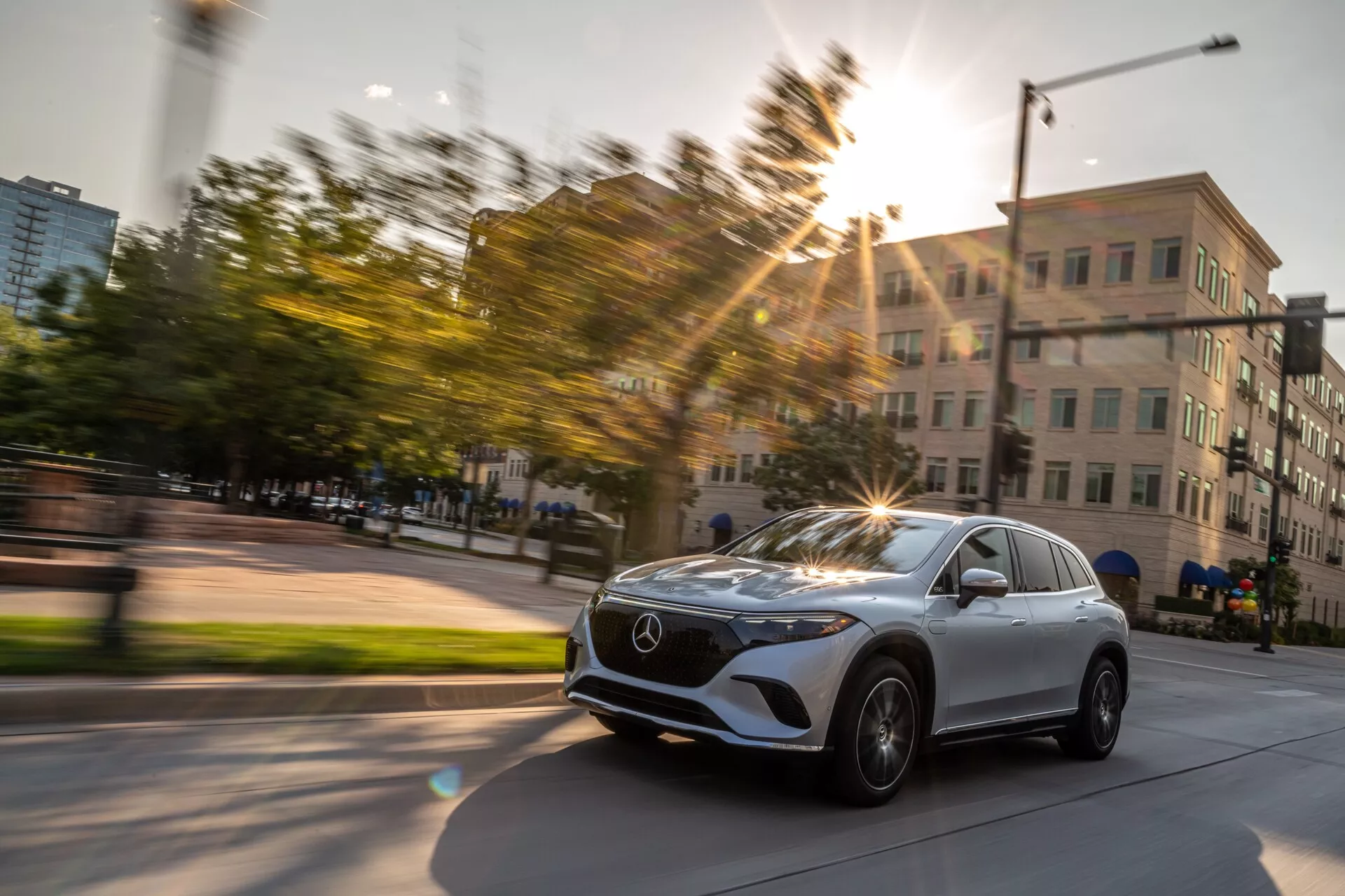 Dynamický snímek vozu Mercedes EQS SUV 2022 zachycený v pohybu z mírně diagonálního předního tříčtvrtečního pohledu. Fotografie zdůrazňuje přední mřížku vozidla s emblémem Mercedes, elegantní LED světlomety, kolo a blatník na straně řidiče a část čelního skla a dveře řidiče. Kola vozu jsou otočená doprava, což naznačuje zatáčení nebo změnu směru. Sluneční světlo vytváří efekt světelného závoje, který dodává fotografii energii, pořízenou v městském prostředí s budovami a zelení, během zlaté hodiny. Rozmazání pohybu v pozadí zdůrazňuje rychlost a pohyb.