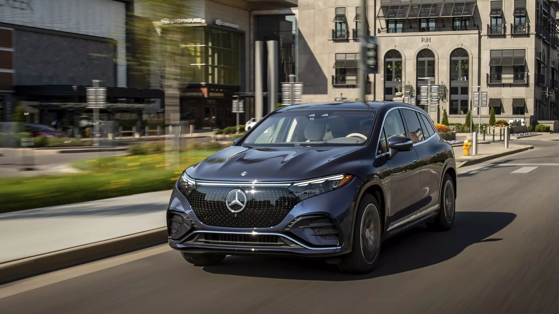 Dynamický snímek SUV Mercedes EQS z roku 2022 v pohybu, zachycený z nízké tříčtvrtinové perspektivy na straně řidiče. Luxusní vozidlo ukazuje svůj výrazný přední gril s velkým emblémem, elegantní LED světla s denním svícením a aerodynamický design. Kola z lehkých slitin a tělově zbarvená boční zrcátka s integrovanými směrovkami zdůrazňují luxus. Městské pozadí s efektem rozmazání pohybu zvýrazňuje pokročilé technologické prvky vozu a jeho přítomnost na silnici.