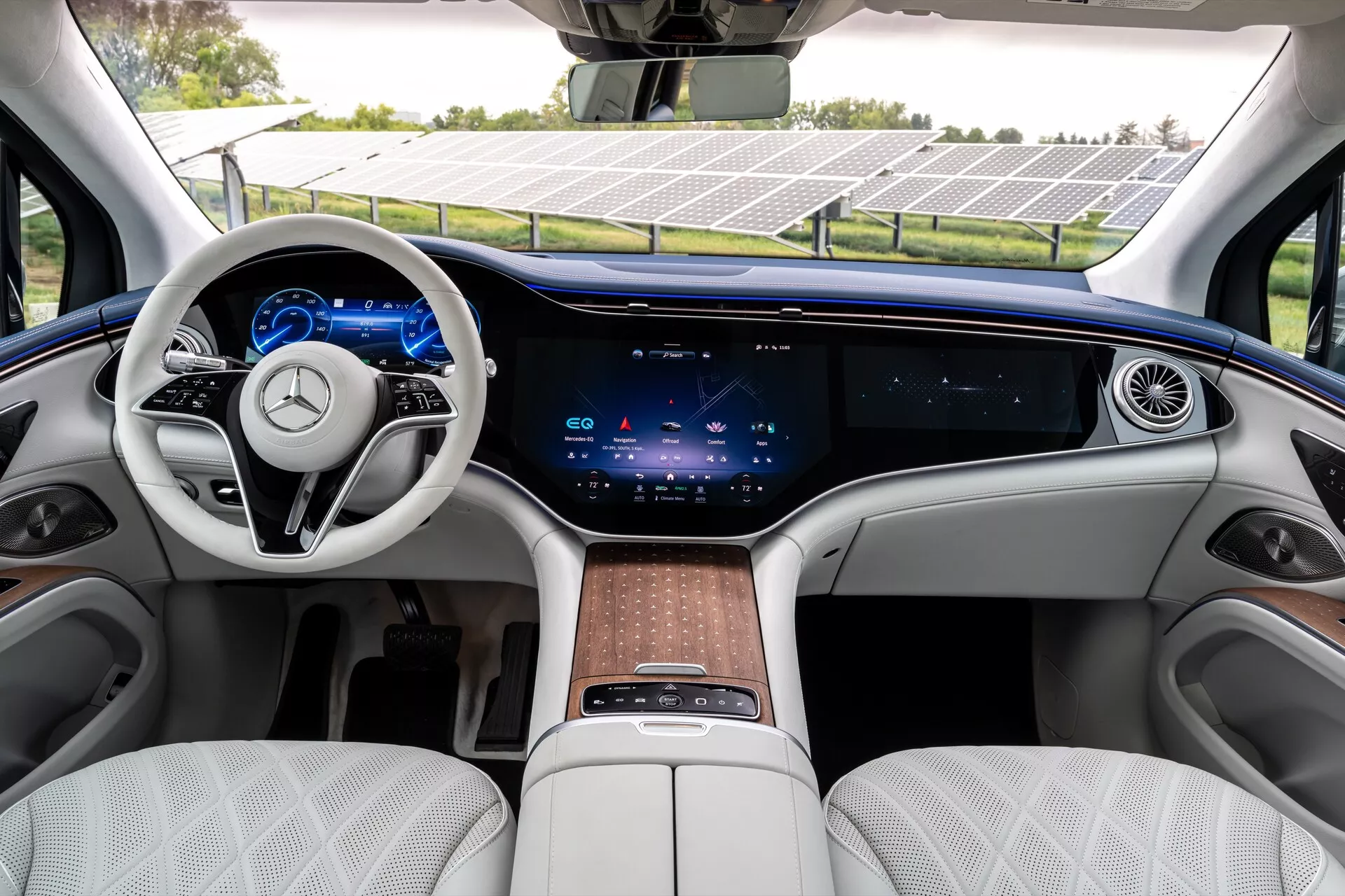 Interiér Mercedesu EQS-SUV 2022, který předvádí luxusní přední řadu s dvouramenným volantem, integrovanými dotykovými ovládacími prvky, plně digitálním přístrojovým panelem, rozlehlým centrálním dotykovým displejem přes celou palubní desku, minimálními fyzickými ovládacími prvky pro klimatizaci, kontinuálním větracím otvorem, bílými prošívanými koženými sedadly, dřevěnými obloženími a centrální konzolí s dotykovou podložkou a úložným prostorem.