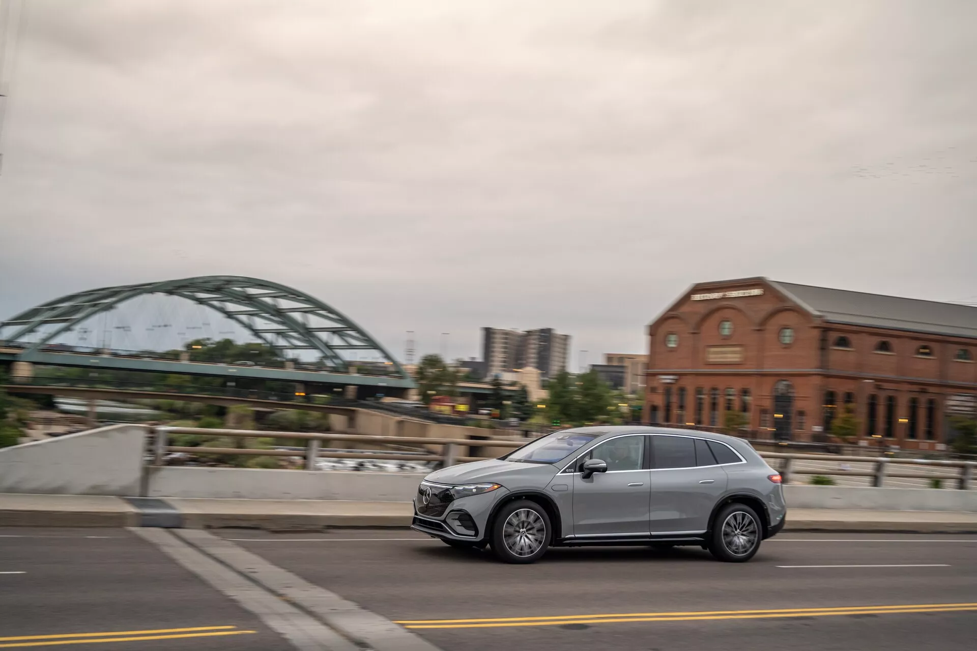 Boční pohled na moderní Mercedes EQS-SUV 2022 jedoucí po městské ulici, ukazující elegantní design, pět dveří, hliníková kola a LED světlomety. Zachyceno z mírně zadního úhlu s rozmazaným pozadím.