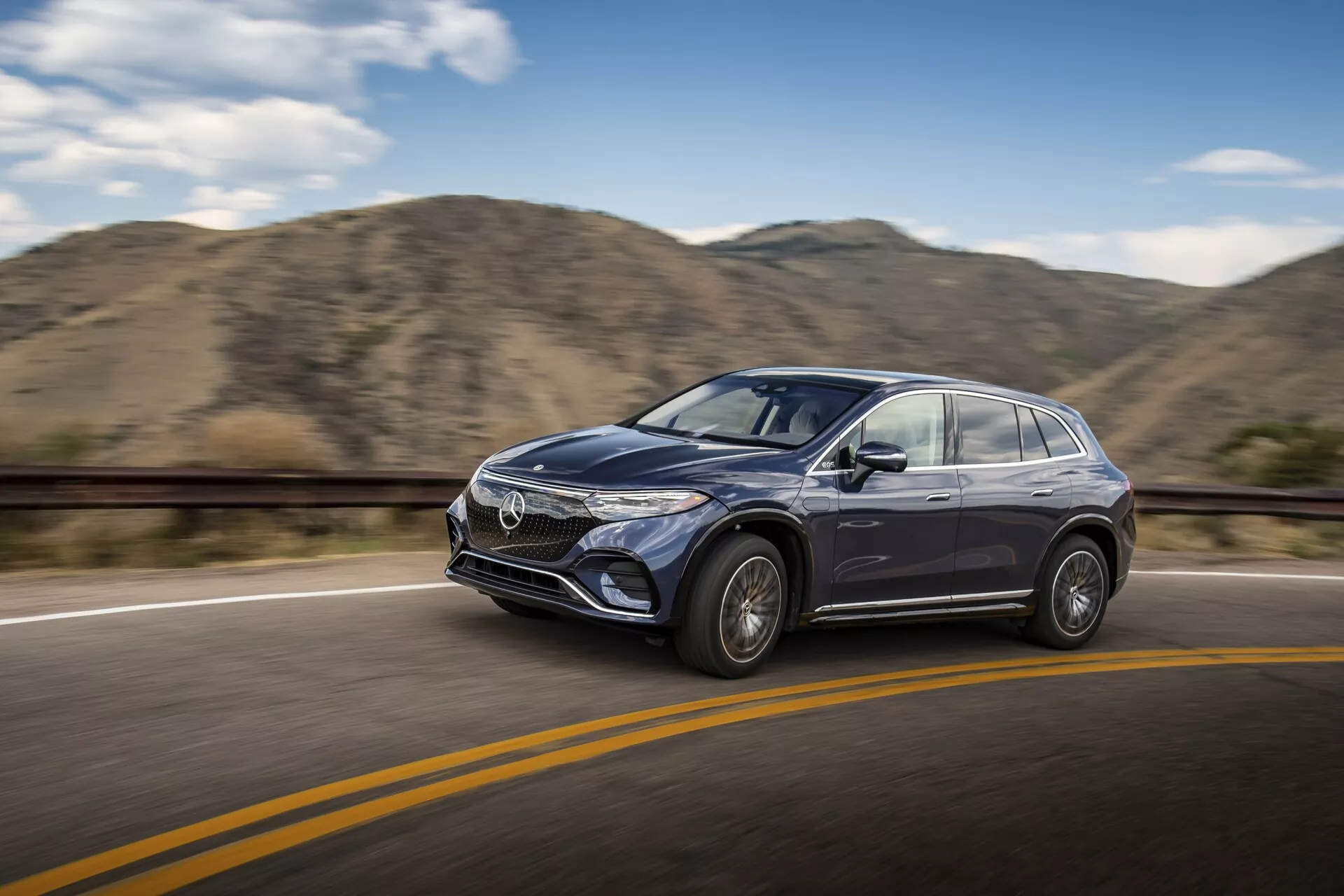 Záběr v pohybu moderního Mercedesu EQS-SUV 2022 jedoucího po hladké asfaltové silnici s žlutou rozdělovací čarou pod jasnou oblohou. Auto je viděno z mírného úhlu přední strany, zdůrazňující řidičovu stranu, včetně přední mřížky, designu světlometů, dveří na straně řidiče, zpětného zrcátka a části čelního skla. Vozidlo je zaostřeno s rozmazaným pozadím s kopci, což naznačuje pohyb. Viditelné jsou designové prvky jako kola z lehkých slitin, tvarované boční panely, výrazné stylové linie a potenciální technologie ADAS.