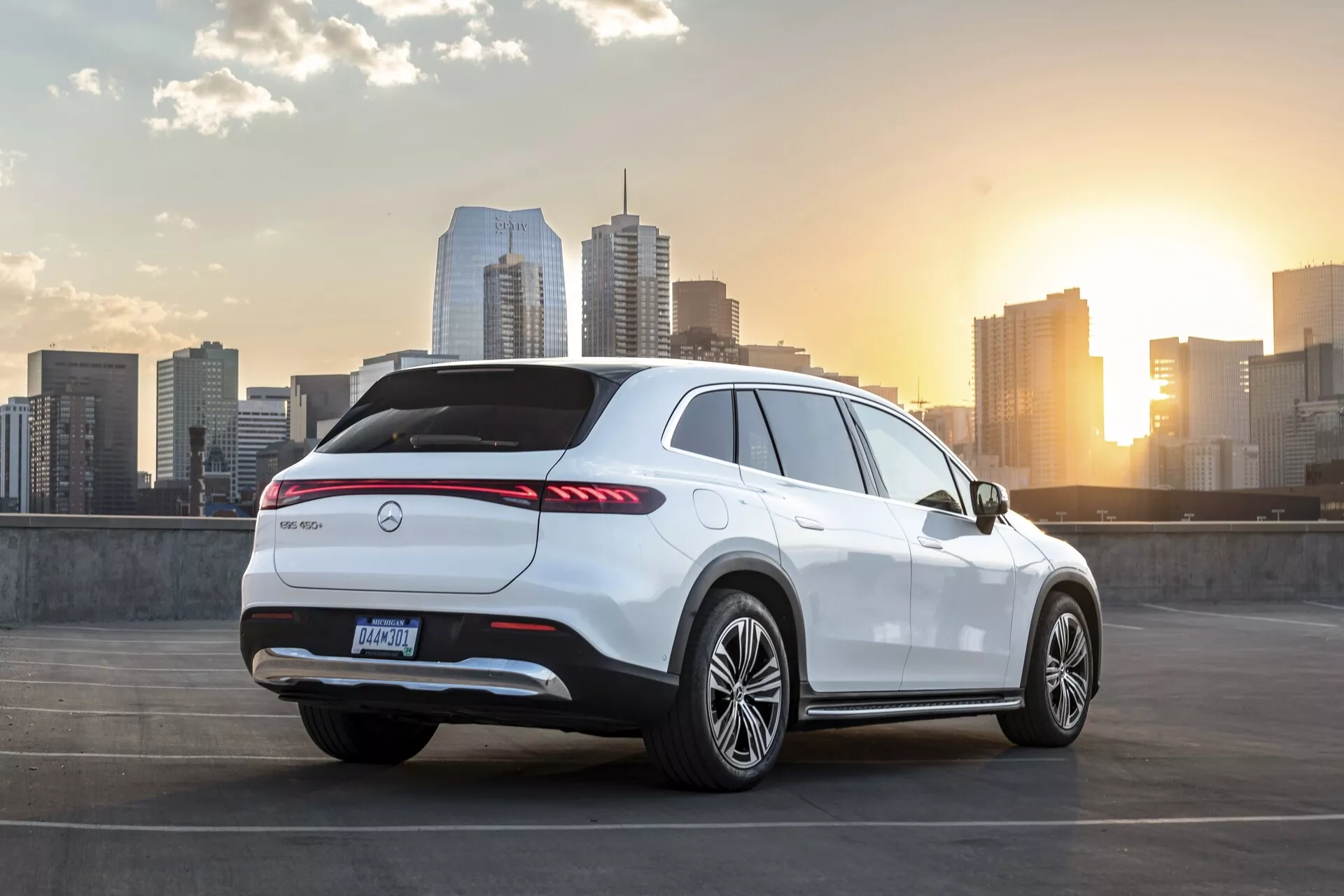 Obrázek bílého SUV Mercedes EQS z roku 2022 zaparkovaného na horní úrovni parkovacího domu. Vozidlo je viděno z třičtvrtinového zadního pohledu, zobrazujícího zadní, boční a část střešní linky. Obsahuje dlouhé zadní LED světlomety, emblém výrobce ve středu zadního víka, zadní difuzor, zadní spoiler, hliníková kola a vysokou linii pásu se sportovně skloněnou střechou. Scéna zahrnuje panoráma města a západ slunce v pozadí, zvýrazňující témata městského luxusu a moderního životního stylu.