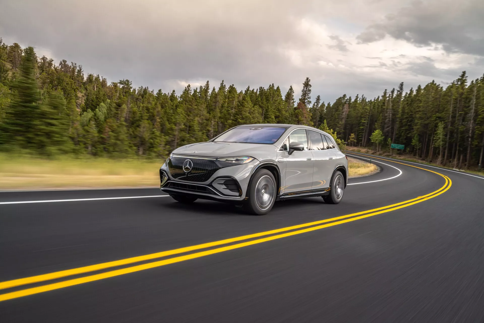 Tento obrázek zachycuje Mercedes-Benz EQS SUV 2022 v pohybu z tříčtvrtečního čelního úhlu s viditelným čelním designem a bočním profilem. Vozidlo je umístěno vlevo, jede po dobře udržované asfaltové silnici s dvojitými žlutými čarami a pravotočivou zatáčkou. Rozmazání kol a mírné rozmazání způsobené pomalejší rychlostí závěrky zdůrazňují rychlost a dynamický pohyb. Zatažené světelné podmínky tvoří měkké stíny a rovnoměrné osvětlení vozidla. Technologické prvky, jako jsou LED světlomety, denní svícení, velká přední maska s emblémem a aerodynamická boční zrcátka s možnými senzory, jsou viditelné. Stříbrný nebo světle šedý lak kontrastuje s lesklou černou spodní částí masky, lemy blatníků a okenními lištami, což zdůrazňuje elegantní linie, zakřivené blatníky a kupé podobnou střechu. Vhodné pro kategorie automobilů, technologie, fotografování v pohybu a luxusní vozidla.