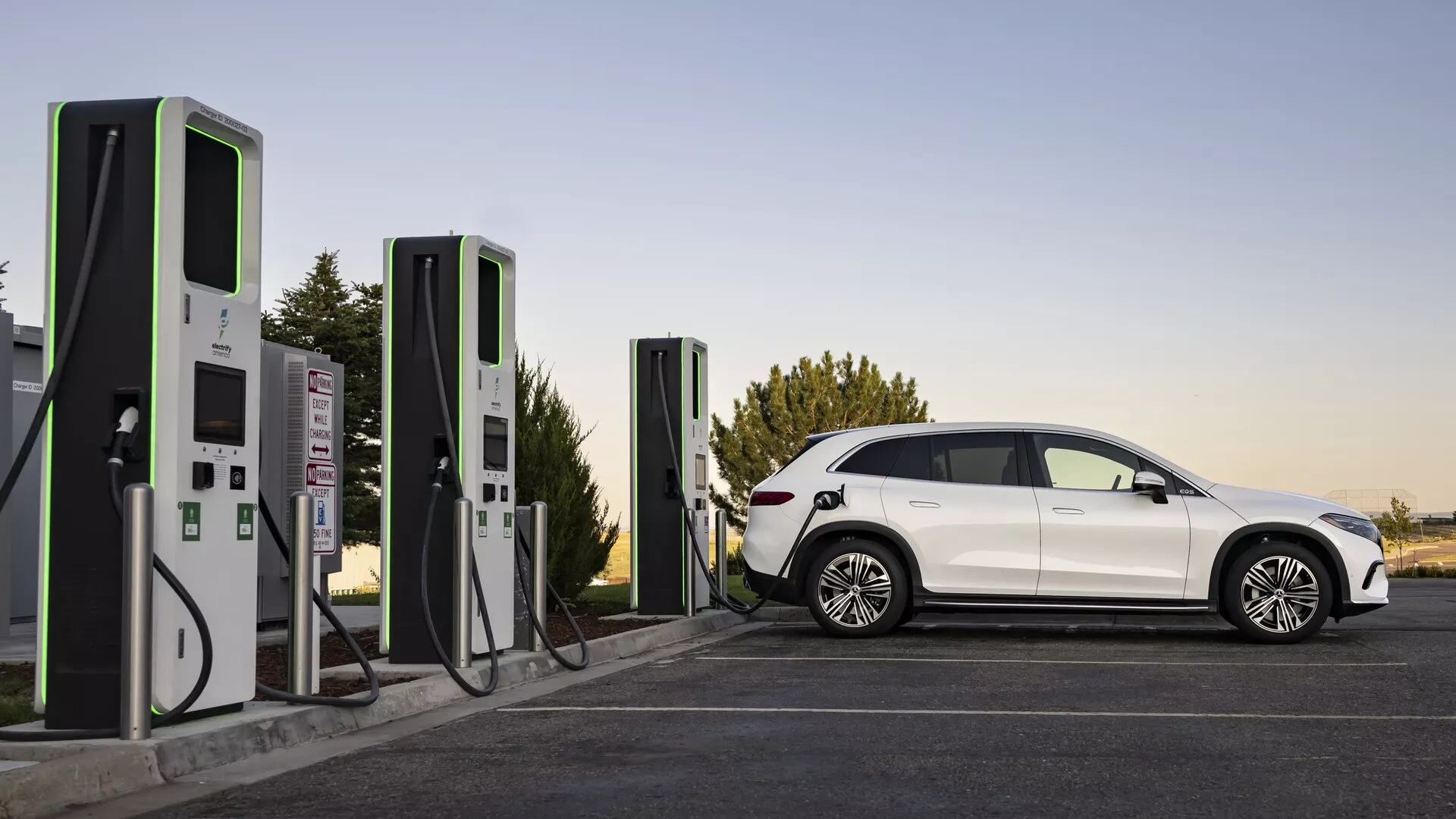 Elegantní a moderní elektrické vozidlo (EV), Mercedes EQS-SUV 2022, ukázané u nabíjecí stanice za měkkých světelných podmínek, buď pozdě odpoledne nebo brzy ráno. Úplný profil bočního pohledu odhaluje aerodynamické linie, výrazná kola, zatmavená okna a dobře definovaný prostor pro cestující. Vozidlo je připojeno k moderní nabíjecí stanici, což zdůrazňuje zaměření na udržitelnou dopravu a infrastrukturu pro elektromobily.
