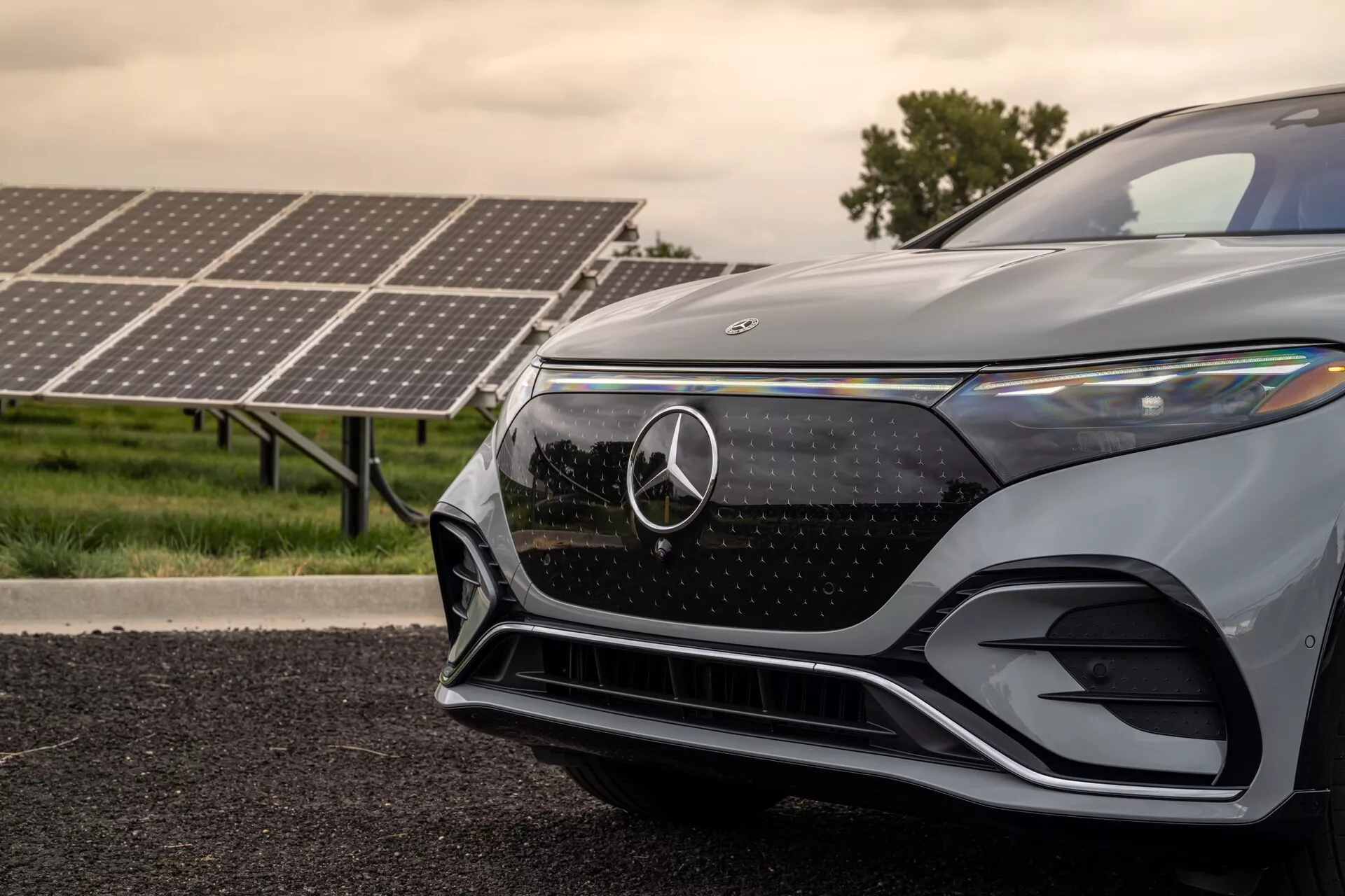 Detailní pohled na přední levý čtvrt Mercedes-Benz EQS SUV 2022 z nízkého úhlu. Obrázek zdůrazňuje mřížku s emblémem Mercedes-Benz, stylový úhlový LED světlomet, aerodynamický přední nárazník a metalický lak. Solární panely na pozadí naznačují elektrickou nebo hybridní povahu vozu a zdůrazňují jeho ekologické a pokročilé technologie.