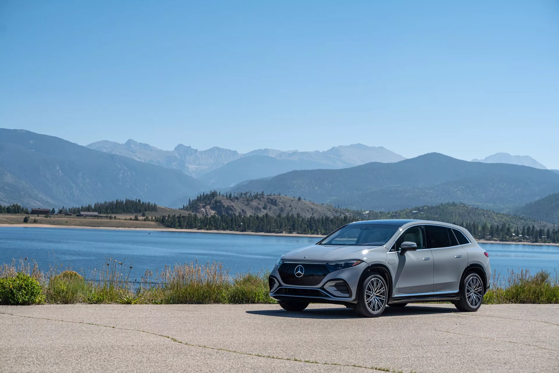 Na obrázku je moderní Mercedes EQS SUV 2022 zaparkovaný na dlážděné ploše s malebným pozadím vody a pohoří, foceno z nízkého úhlu pro zdůraznění jeho postoje a designových linií. Auto je viděno z bočního profilu s viditelnou přední maskou, hranatými LED světlomety, velkými slitými koly, aerodynamickými bočními zrcátky a metalickým lakem.