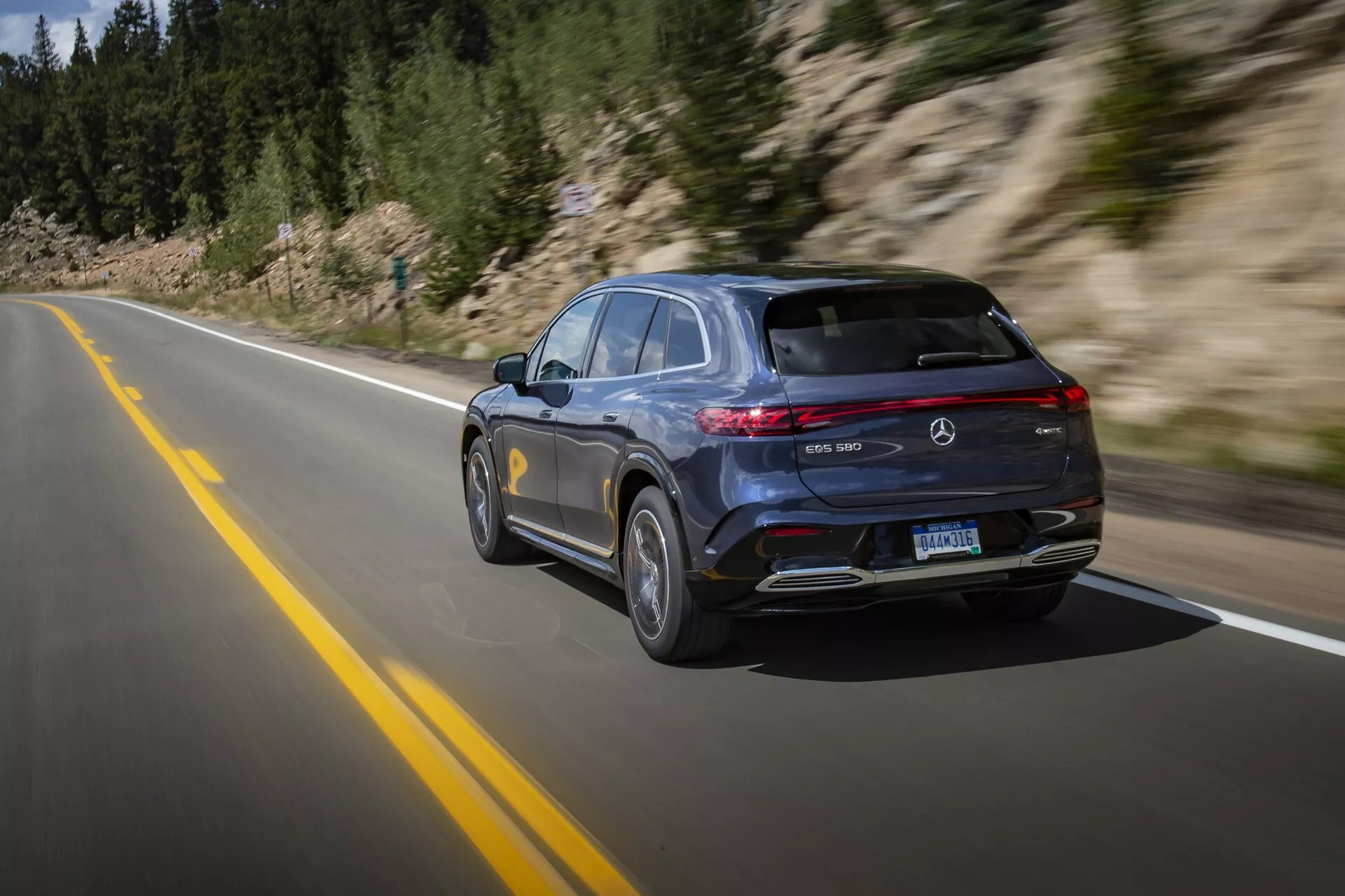 Moderní, luxusní Mercedes-Benz EQS SUV 2022 zachycený v pohybu na dlážděné silnici, ukazující elegantní design s tmavým lakem, vodorovně orientovanými LED zadními světly, střešním spoilerem, dvěma výfuky a koly z lehkých slitin s mnoha paprsky. Technika panningu ve vysoké rychlosti zdůrazňuje pohyb vozidla přírodním, horským prostředím.