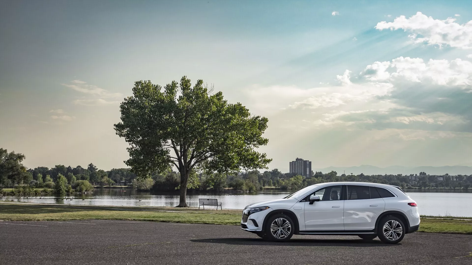 Boční profilní pohled na moderní Mercedes EQS SUV 2022 zaparkovaný na asfaltu, s vícepaprskovými litými koly, elegantními aerodynamickými liniemi, čtyřmi dveřmi, zatmavenými okny s integrovaným ukazatelem směru, sportovní linií střechy s nosníky na střeše a částečně viditelnými LED zadními světly, zasazený do klidného panoramatického pozadí s jezerem nebo řekou, stromem, trávou a částečně zamračenou oblohou.